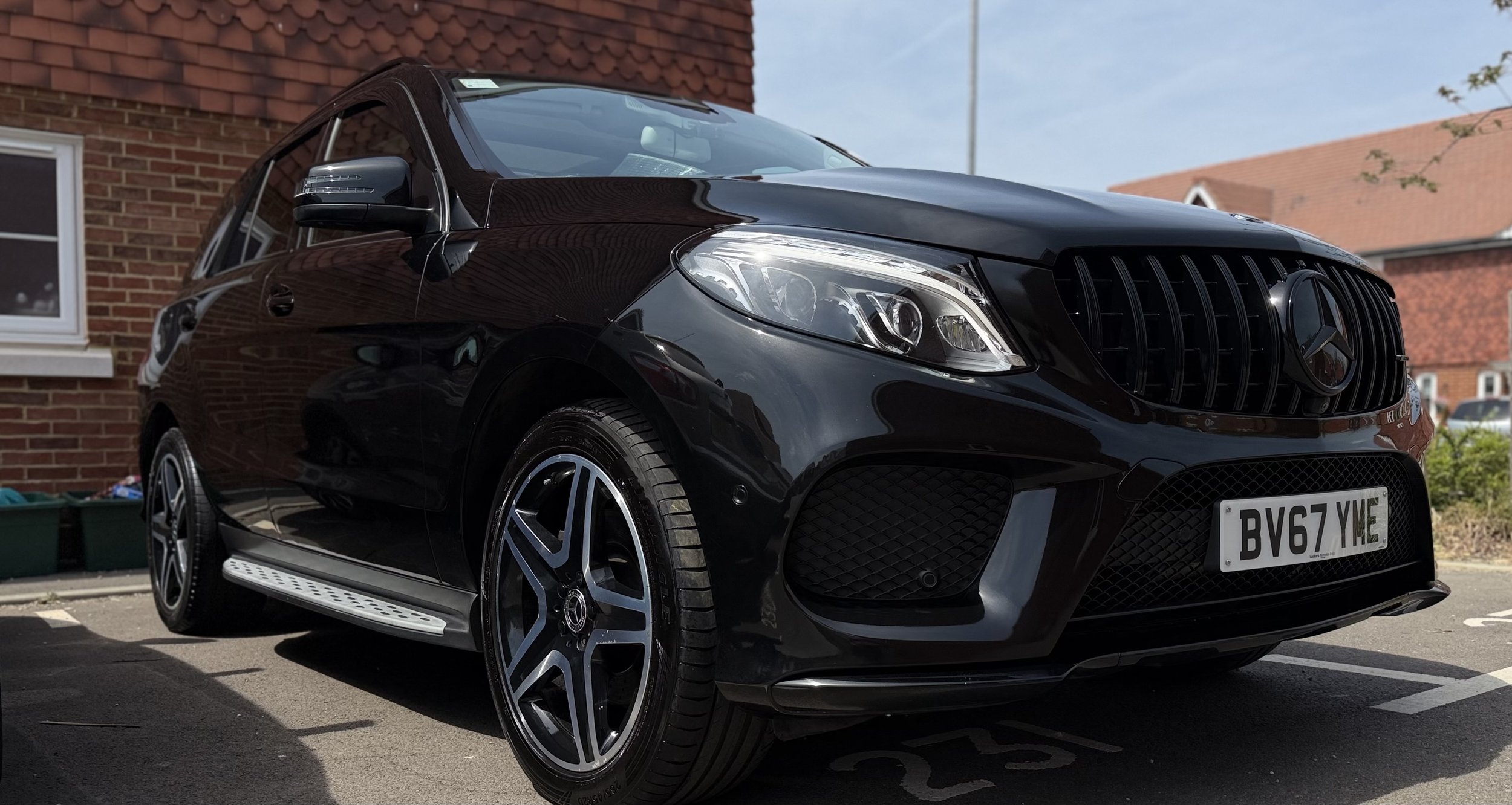 A black Mercedes-Benz SUV parked outdoors in front of a brick house with red roof tiles, under a partly cloudy sky.