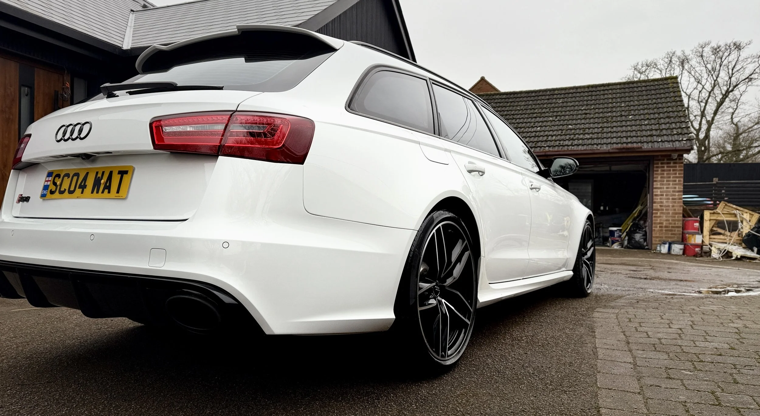 White Audi station wagon parked in front of a garage with a brick wall and tools in the background.