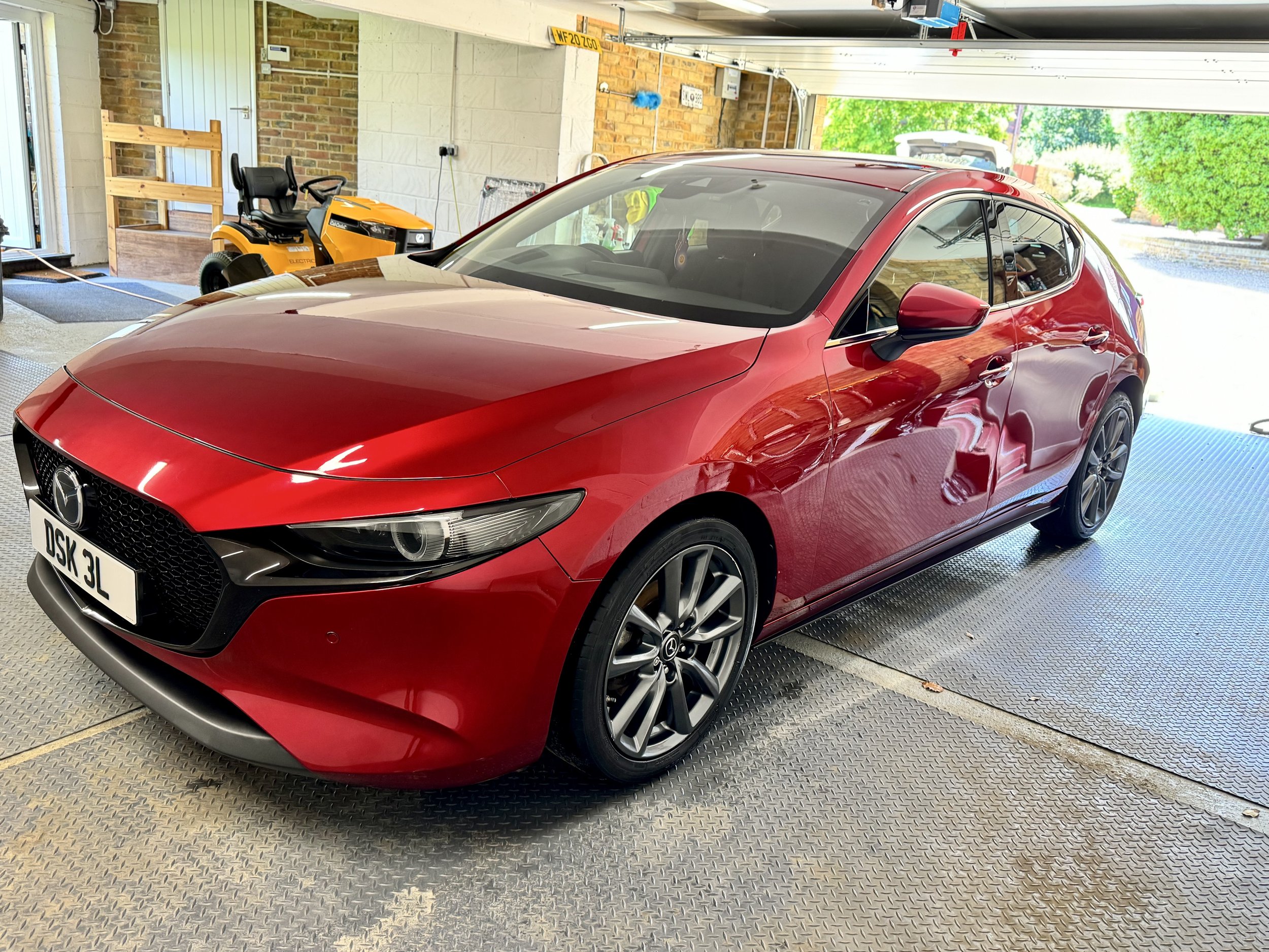 a red Mazda car parked inside a garage with a yellow riding lawn mower in the background and a brick exterior wall visible through the open garage door.