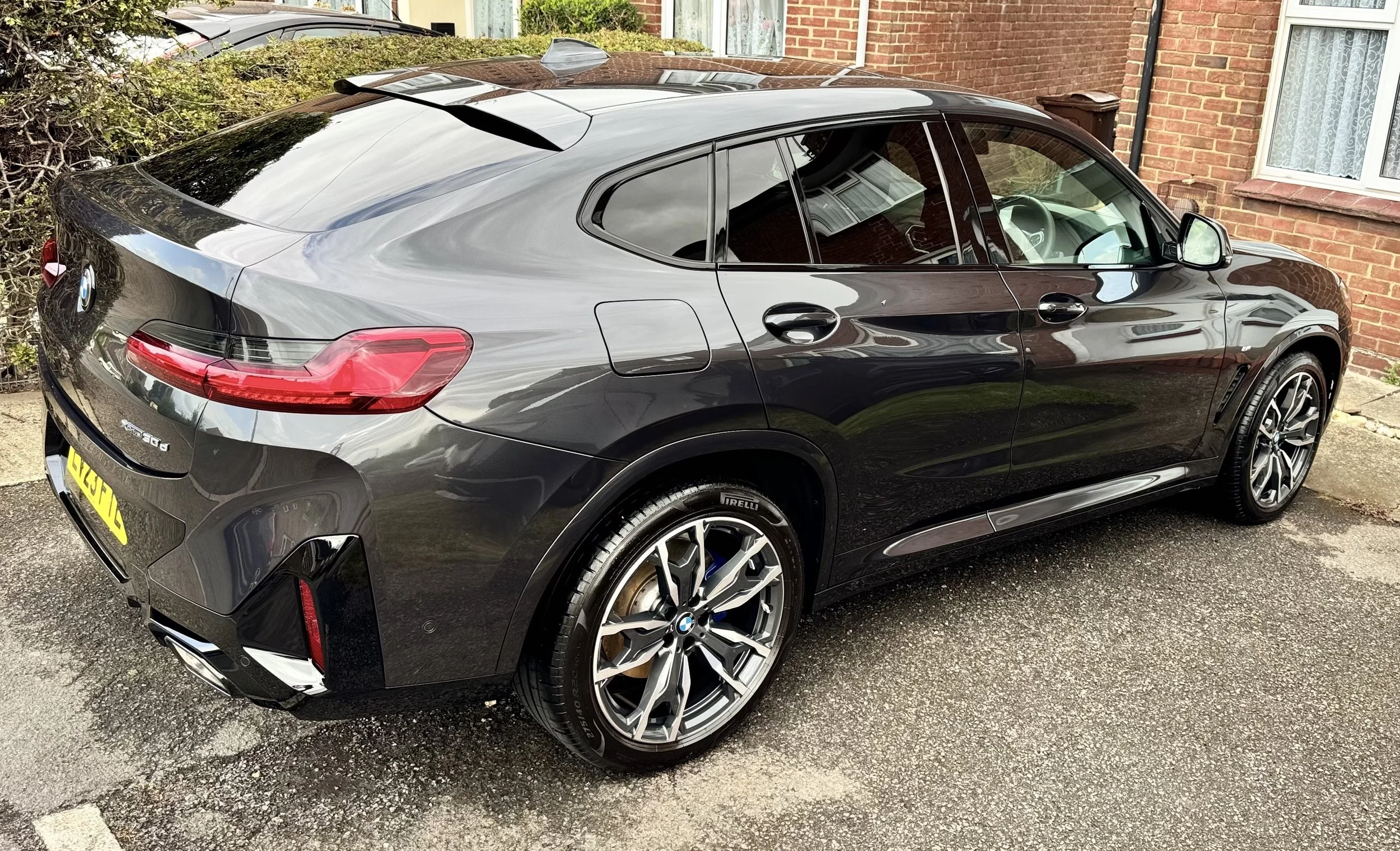 A black BMW coupe parked on a residential driveway in front of a brick house with a window and a bush.