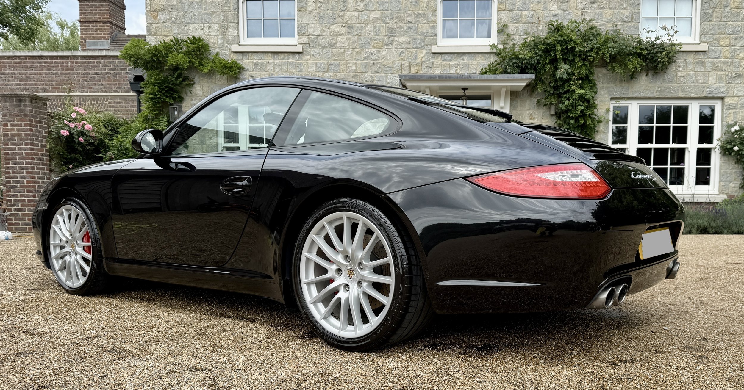 A black Porsche Carrera parked in front of a stone house with white window frames, surrounded by greenery and flowers.
