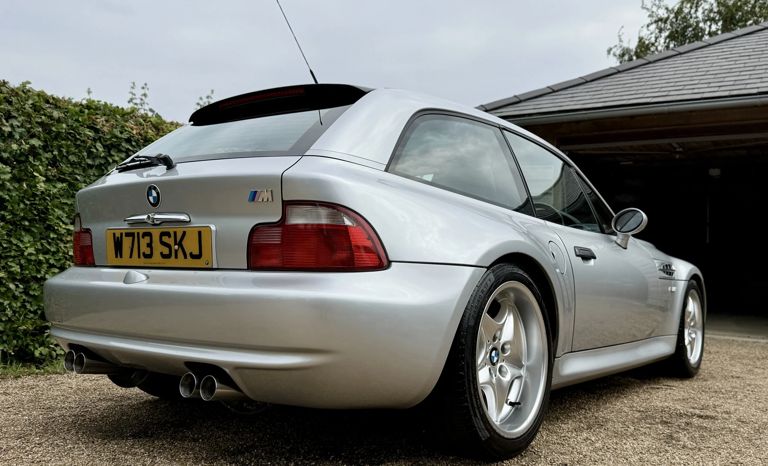 Silver BMW M car parked outdoors, showing the rear and side view with the license plate 'W713 SKJ', surrounded by greenery and a house with a brown roof.