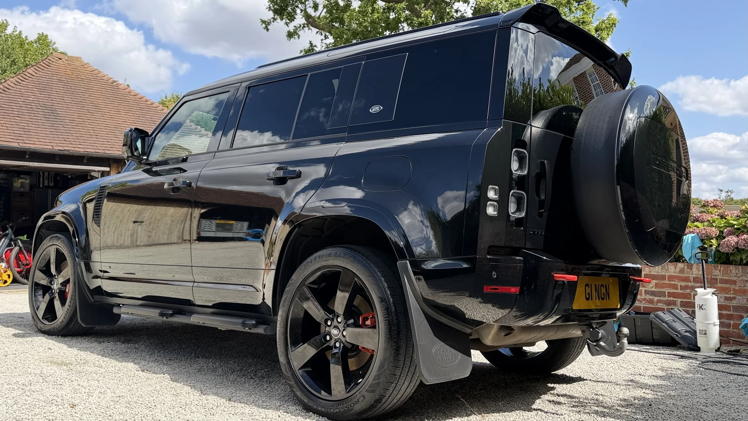 A black Land Rover Defender parked in a driveway, with a spare tire mounted on the rear, a yellow license plate reading 'GI NGN,' surrounded by a garden with flowers, a brick wall, and a house with a garage and red roof in the background on a sunny day with blue skies and some cloud
