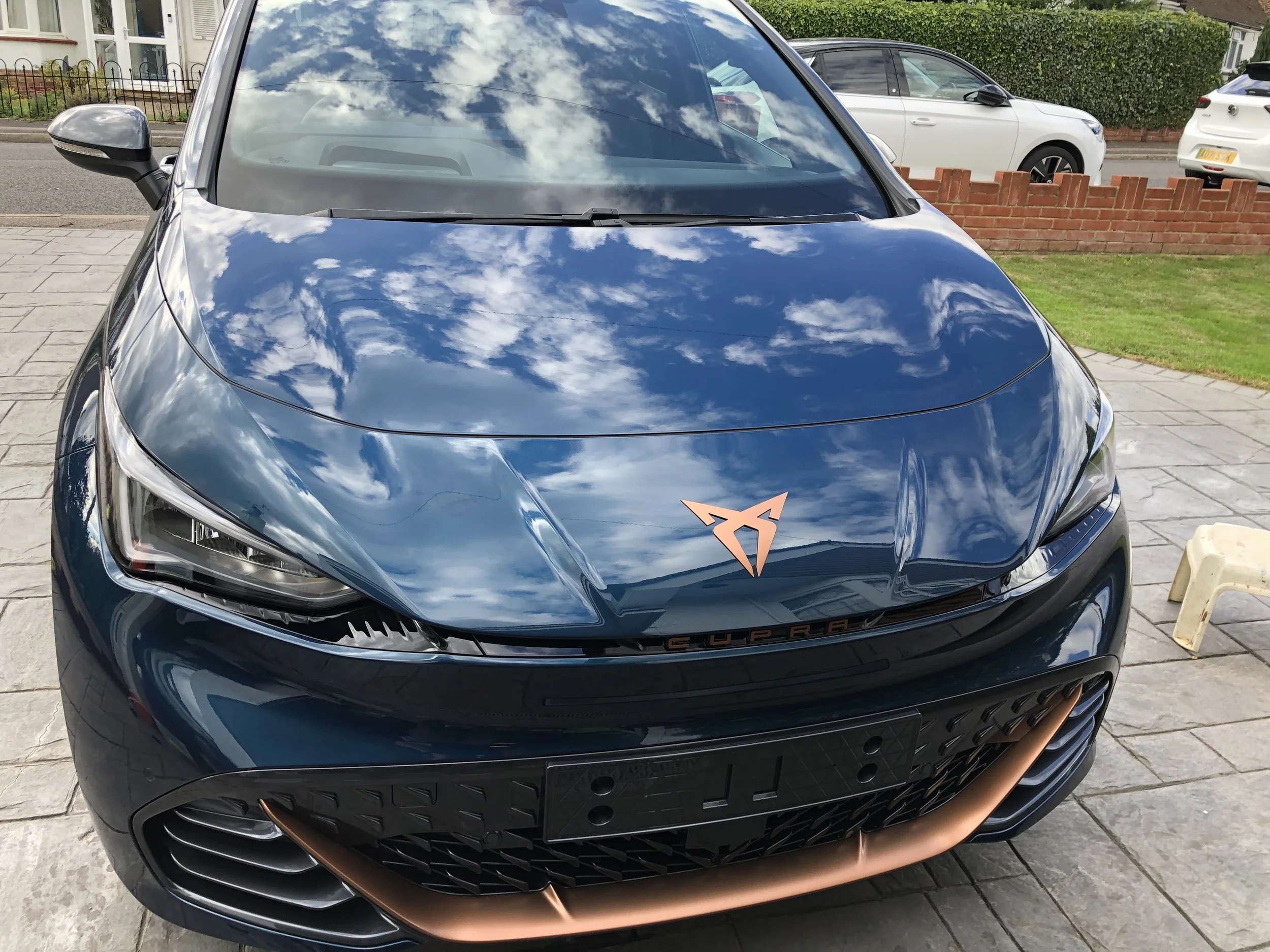 Front view of a dark blue CUPRA car parked on a driveway, with reflections of clouds and sky on the hood.