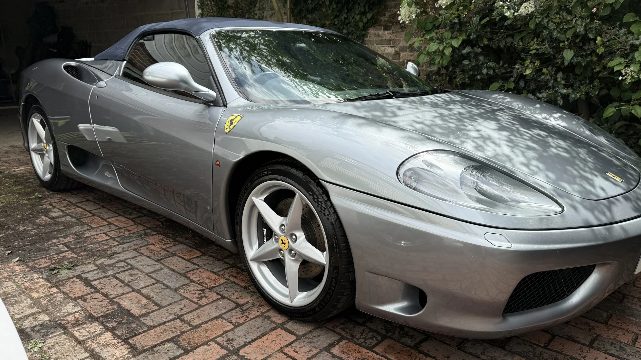 Silver Ferrari sports car parked on brick driveway, surrounded by greenery.