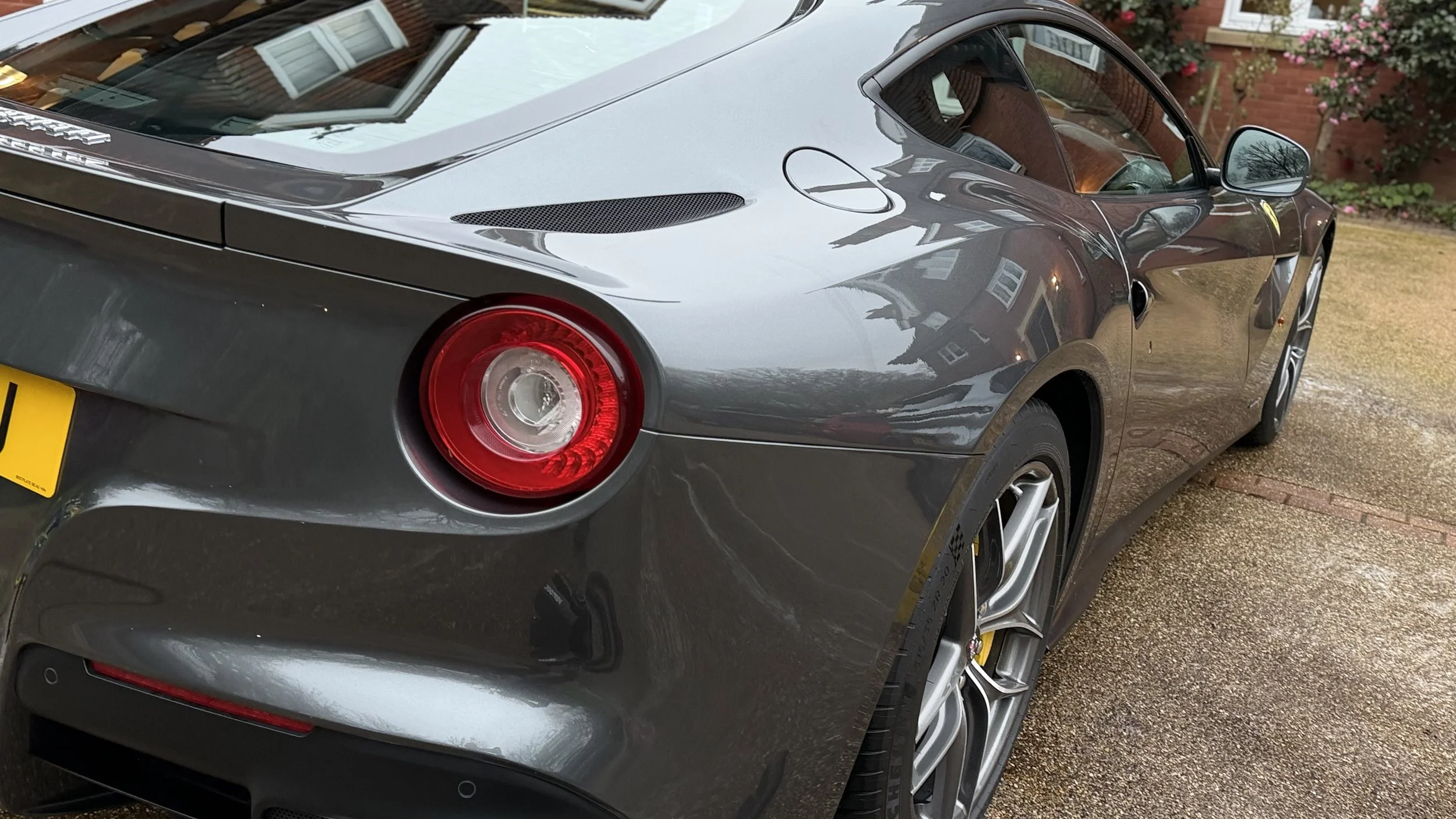 A gray sports car parked on a driveway, with a rear view showing circular taillights, reflections of nearby houses and trees on the car's surface, and a garage door reflected in the windows.
