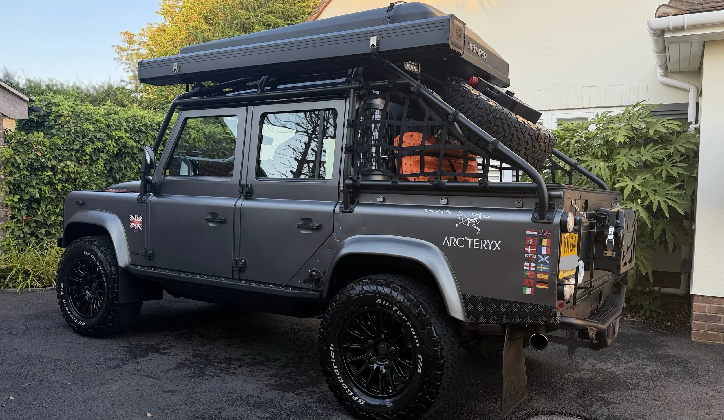 A dark gray off-road vehicle with ARCTERYX logo, supporting flags of various countries, and a UK license plate. The vehicle is equipped with a roof tent, spare tire, orange recovery gear, and a black cargo basket on top. It is parked outdoors near greenery and a white house.