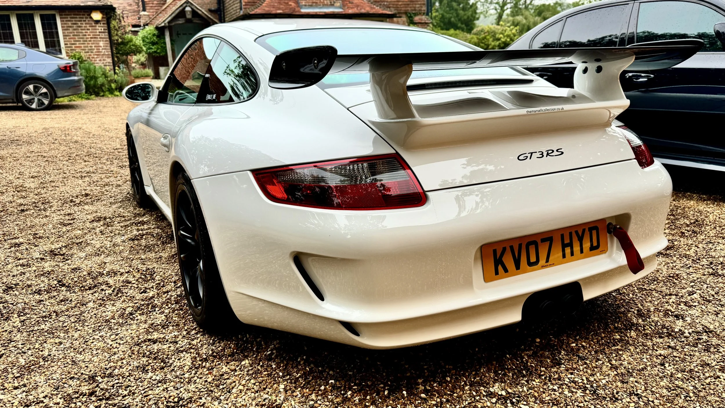 A white Porsche 911 GT3 RS sports car with a large rear wing parked on a gravel driveway, with other cars and a house in the background.