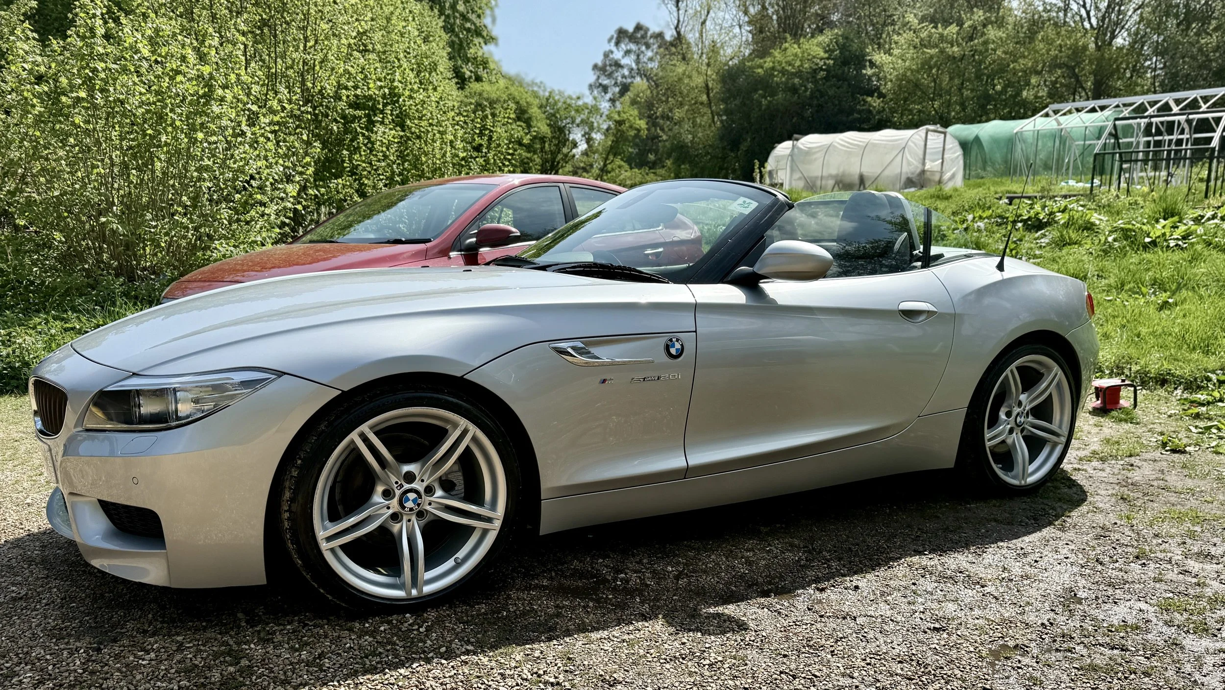 A silver BMW convertible parked outdoors next to a red car, with green trees and garden structures in the background.