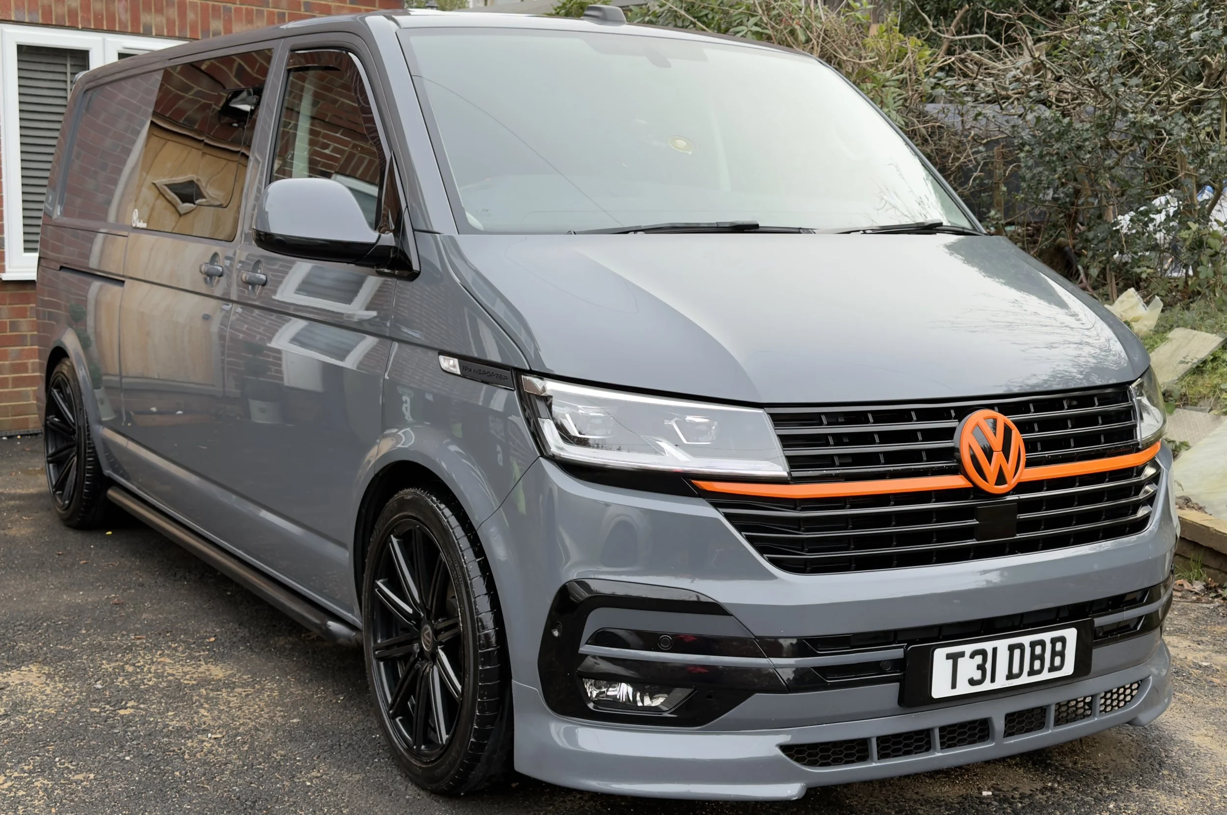 Gray Volkswagen van with custom black wheels and an orange VW logo on the front grille, parked on a driveway near a brick house and bushes.