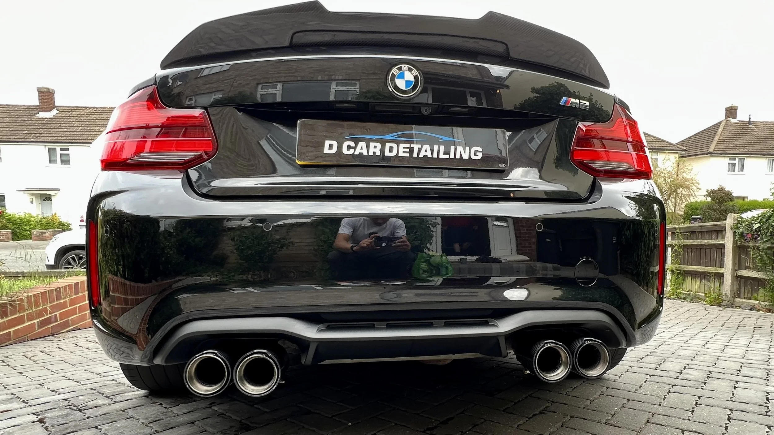 Black BMW M2 with quad exhaust pipes, parked on a brick driveway, reflecting the person taking a photo. Behind it, residential houses and a fence are visible.