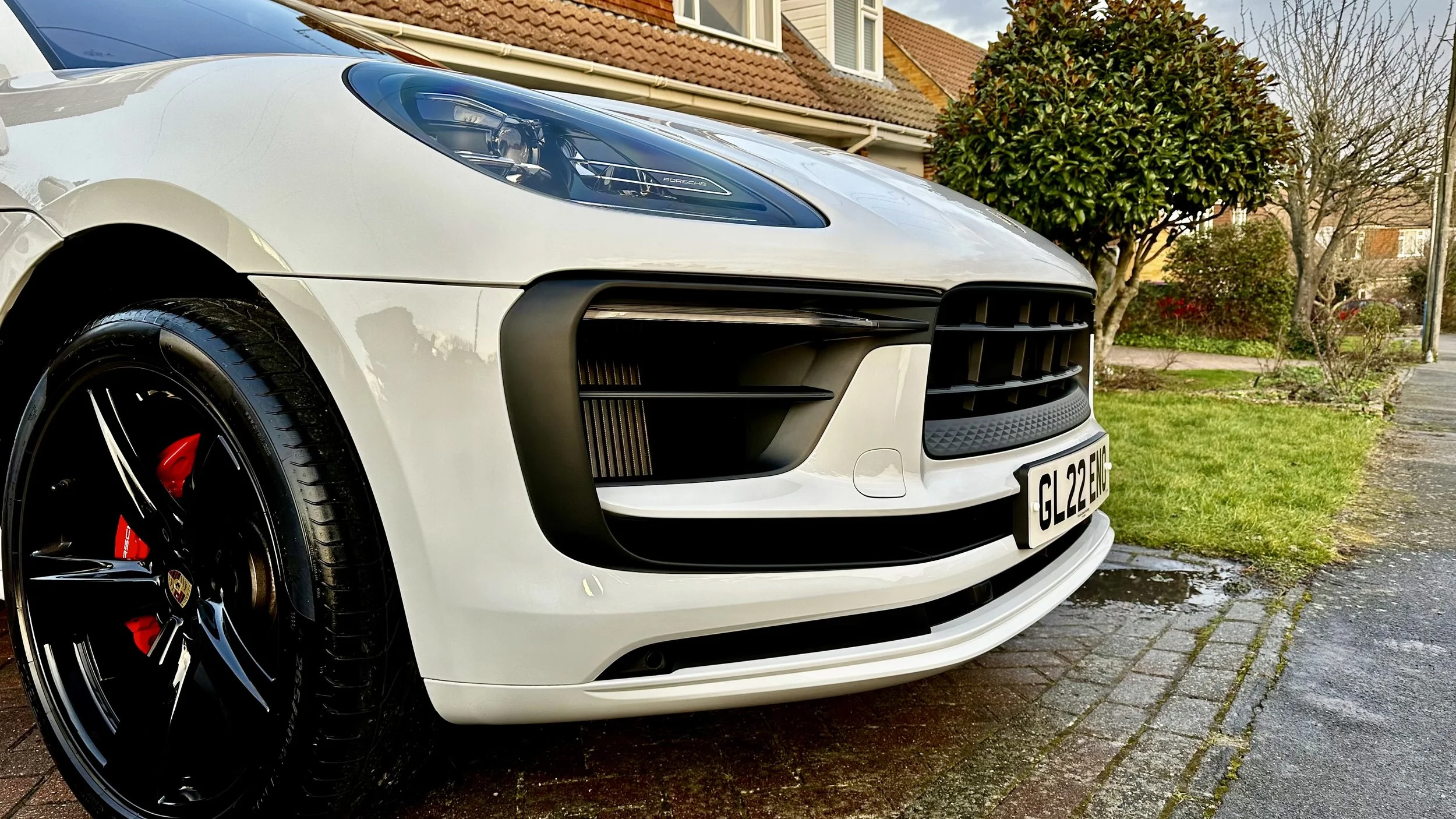 Close-up of the front of a white Porsche car parked on a residential street with a front license plate reading GL22 ENJ, black and red brake calipers visible through the black alloy wheels, and a yard with trees and houses in the background.