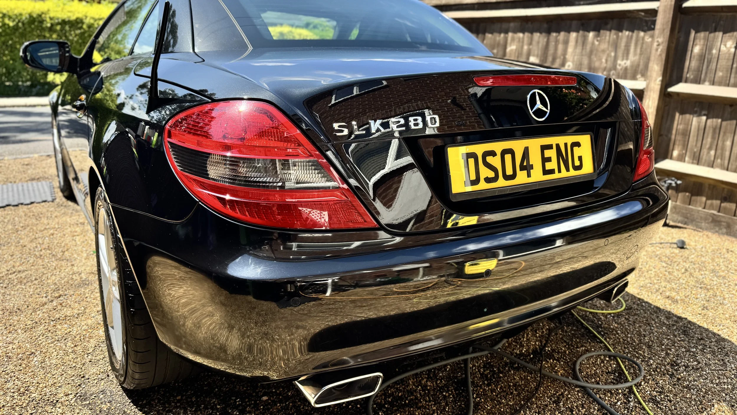 Black Mercedes-Benz SLK280 parked outdoors with a yellow license plate DS04 ENG, mudflaps, rear taillights, and Mercedes logo, reflecting nearby greenery and wooden fence.