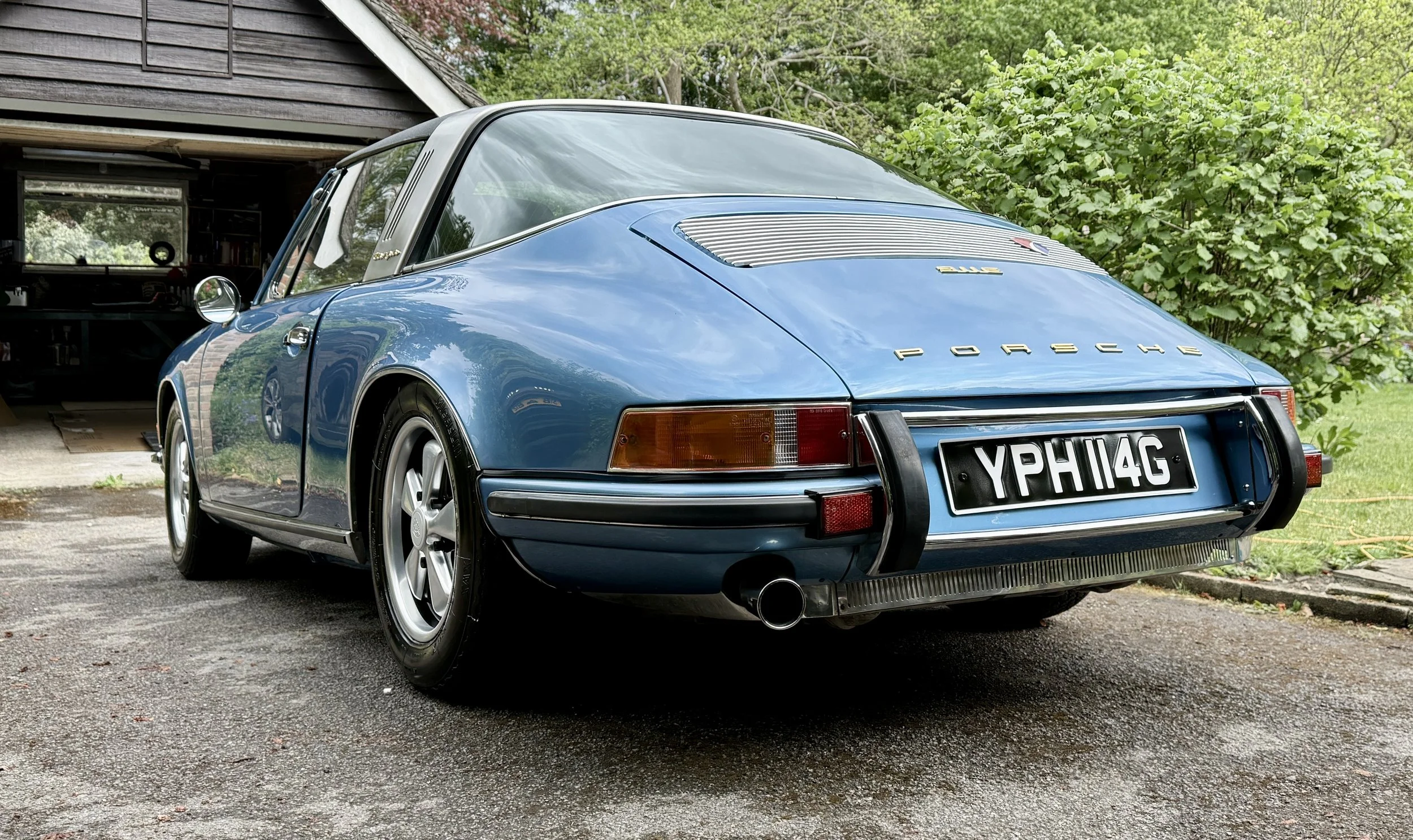 A vintage blue Porsche 911 parked outdoors near a garage and green trees.