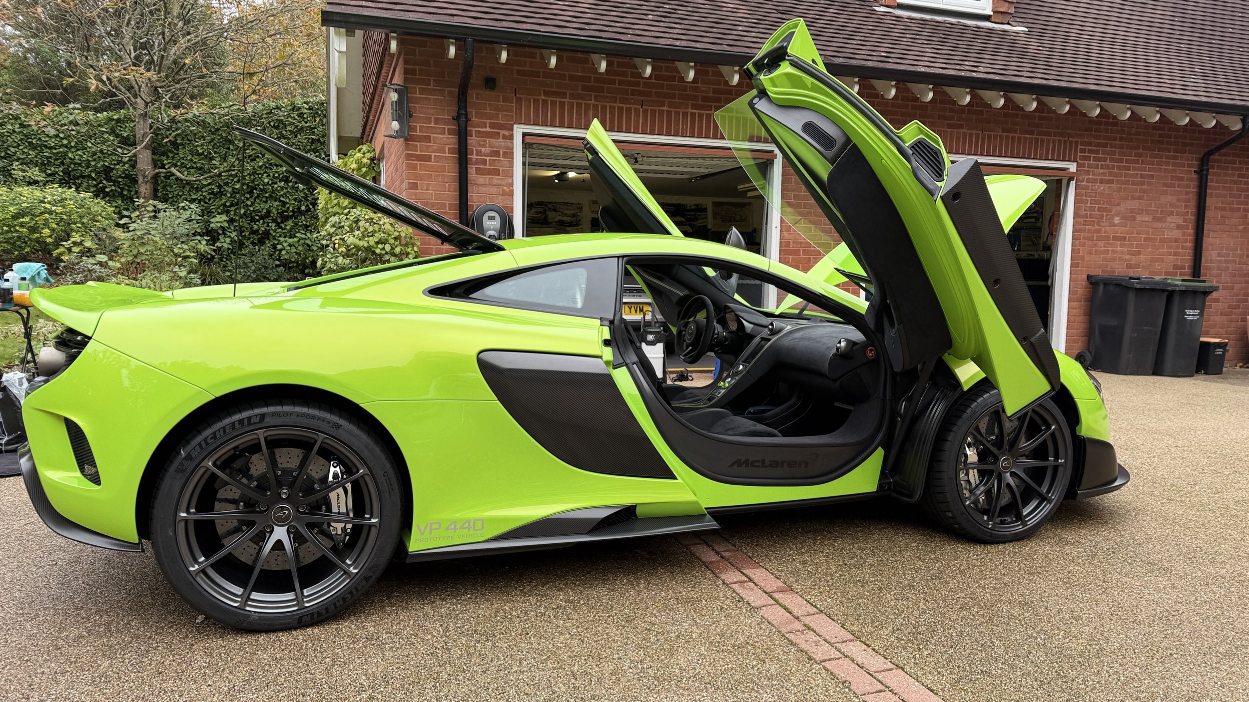 A bright green McLaren sports car with its doors open, parked in front of a brick house with an open garage.