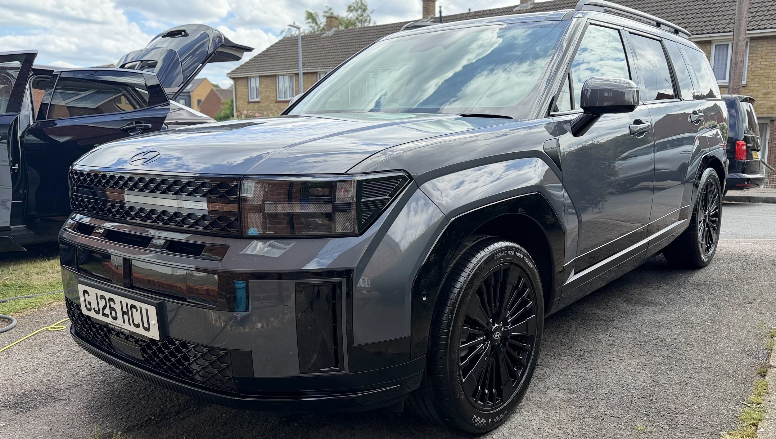 A gray Hyundai SUV with black rims parked on a driveway with houses in the background. The front grille and headlights are visible, and the hood is slightly open.