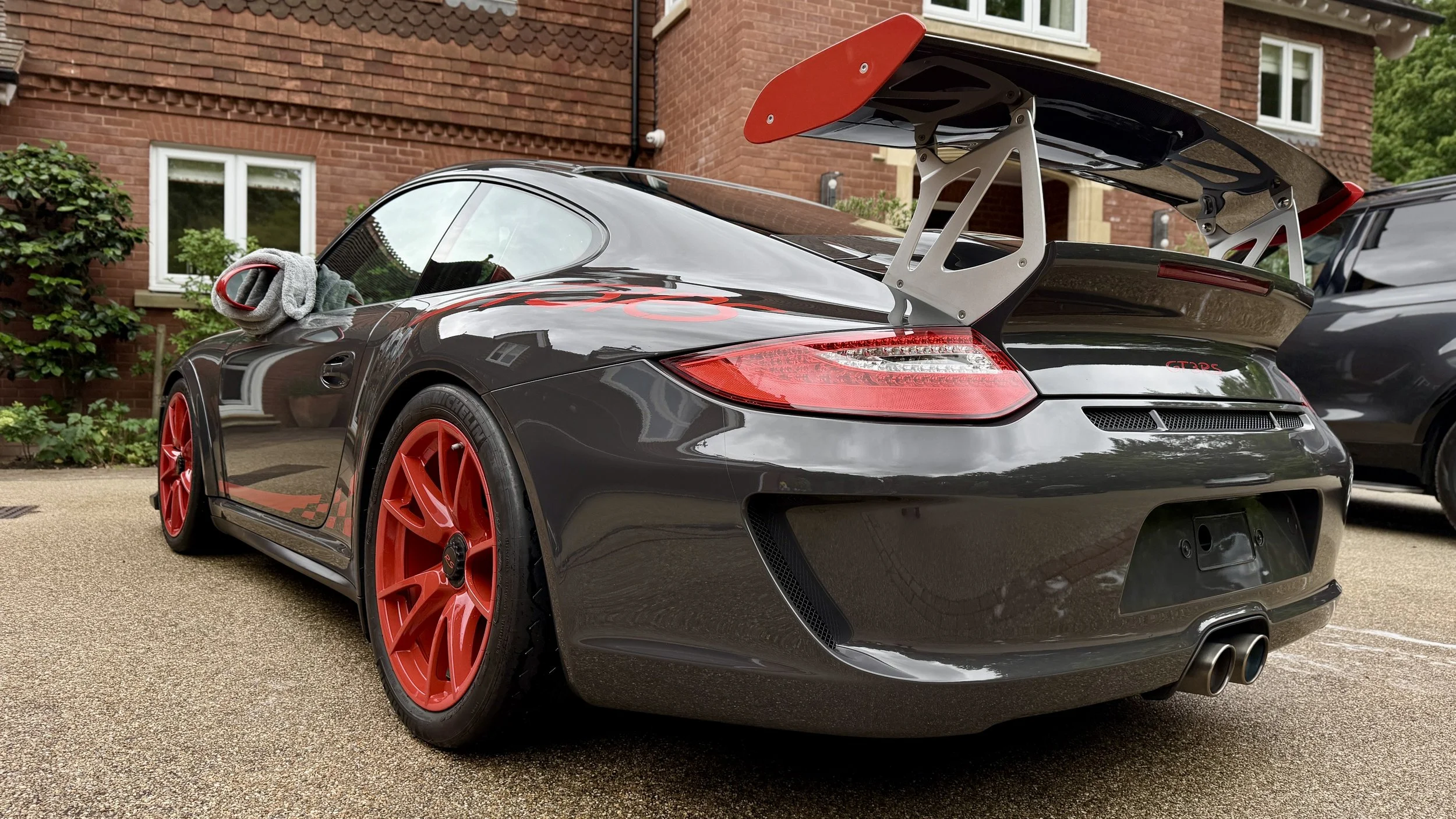 A black sports car with red rims and a large rear spoiler parked outside a brick house.