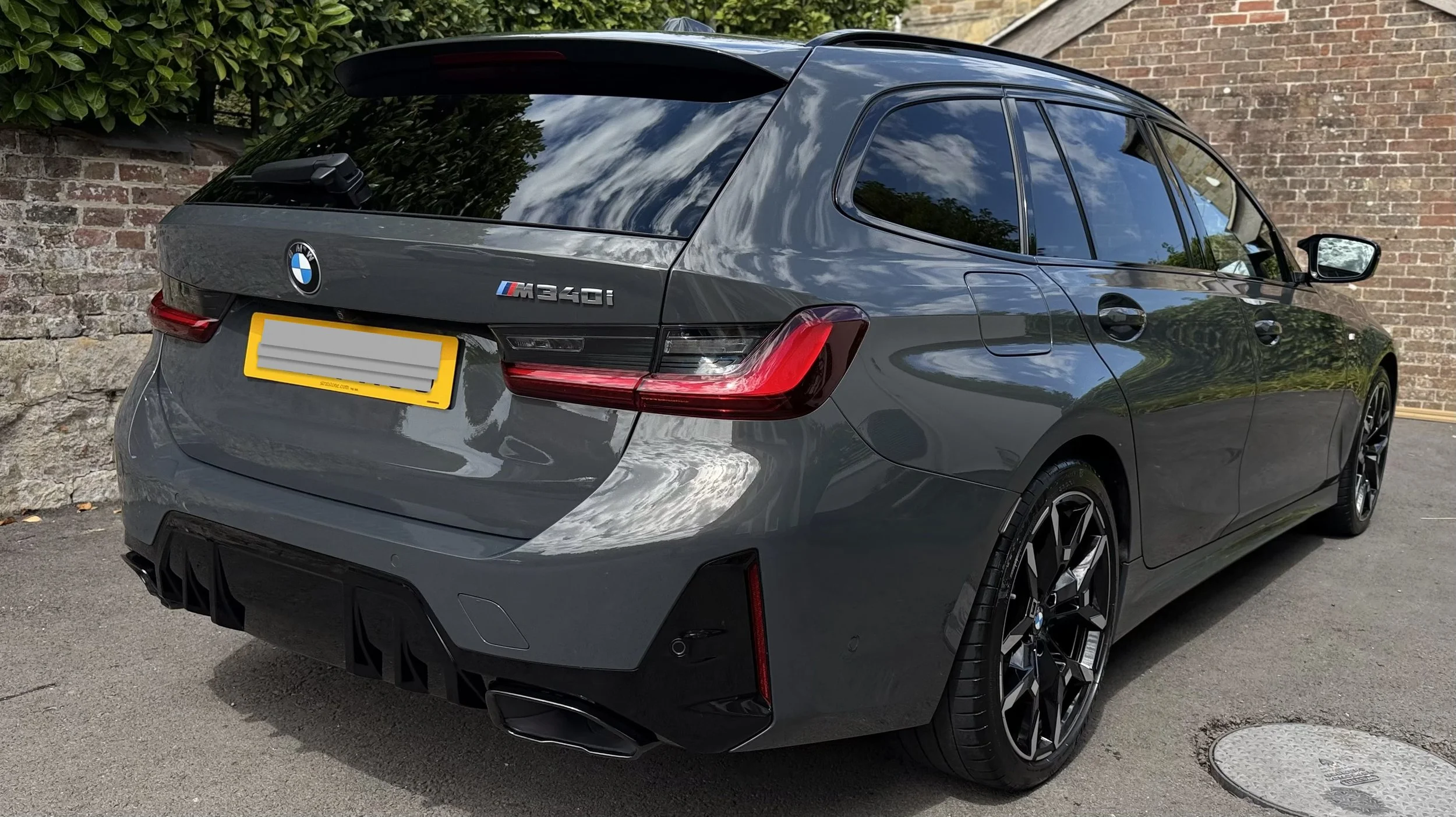 A gray BMW M340i car parked next to a brick wall and greenery on a sunny day, with clouds reflected in the windows.