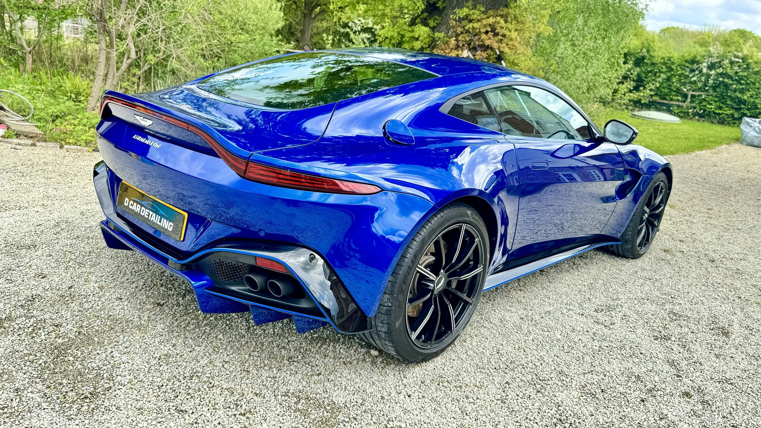 A blue Aston Martin sports car parked on gravel with greenery and trees in the background.