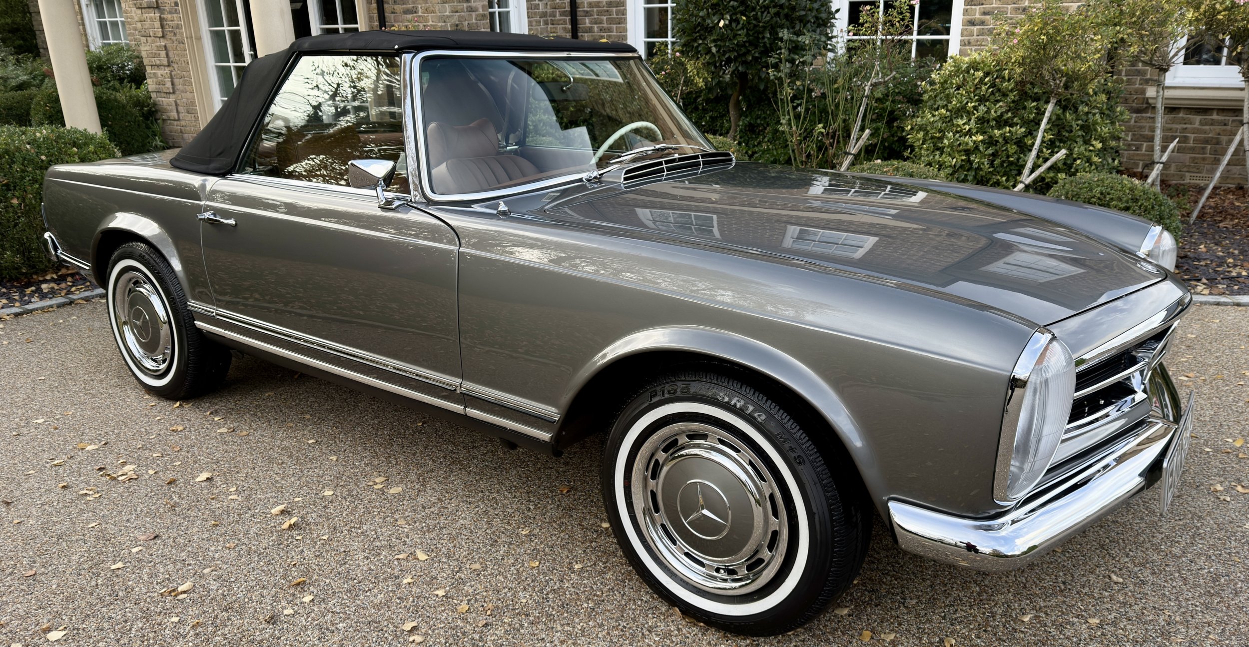 A vintage silver Mercedes-Benz convertible car parked on a driveway with houses and greenery in the background.