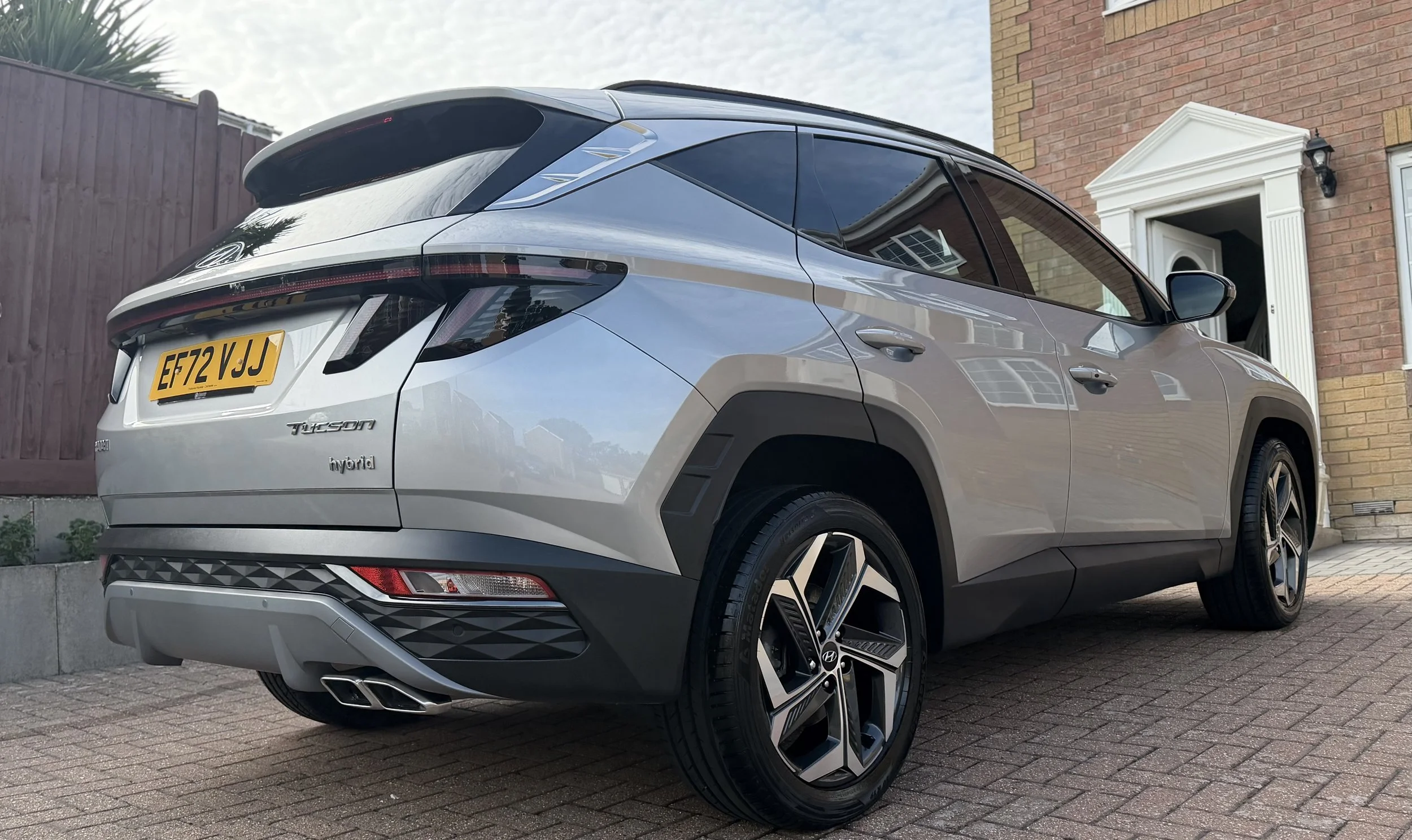 A silver Hyundai Tucson hybrid SUV parked on a brick driveway in front of a brick house with a white door frame and black outdoor light, with a wooden fence and cloudy sky in the background.