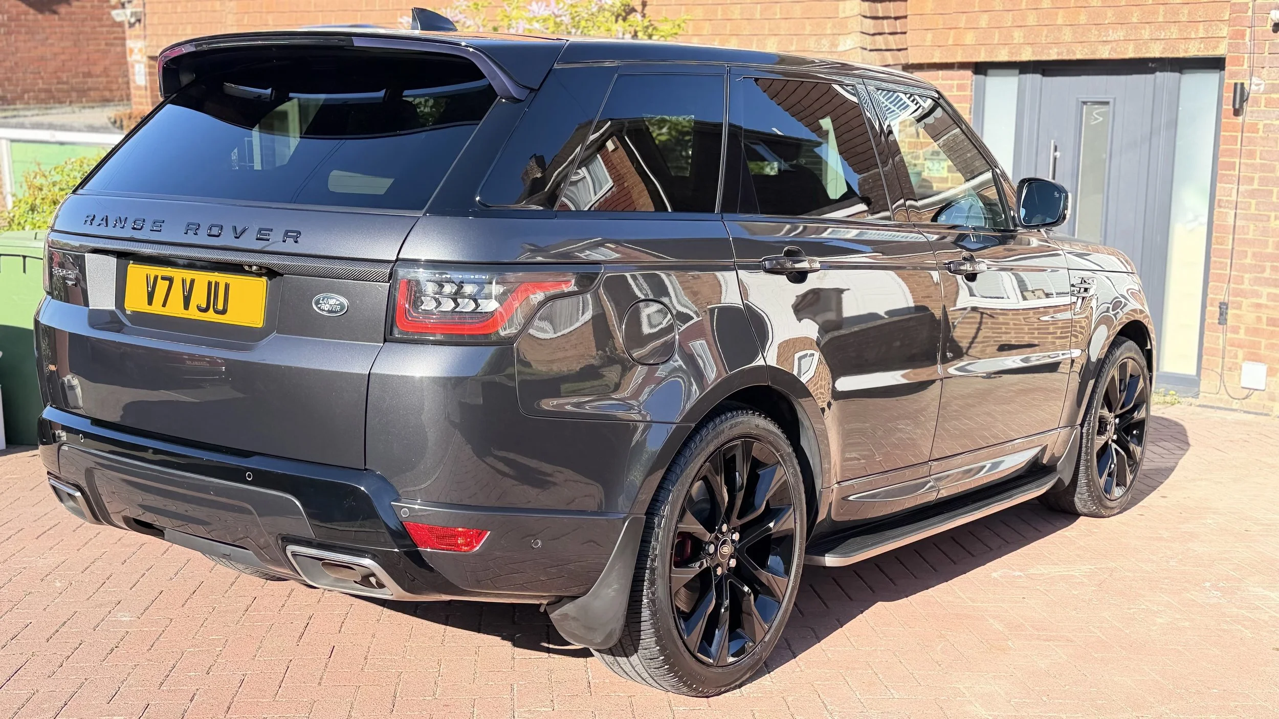 Black Range Rover SUV parked on a brick driveway in front of a brick house with a gray door and window, reflections visible on the car's surface.