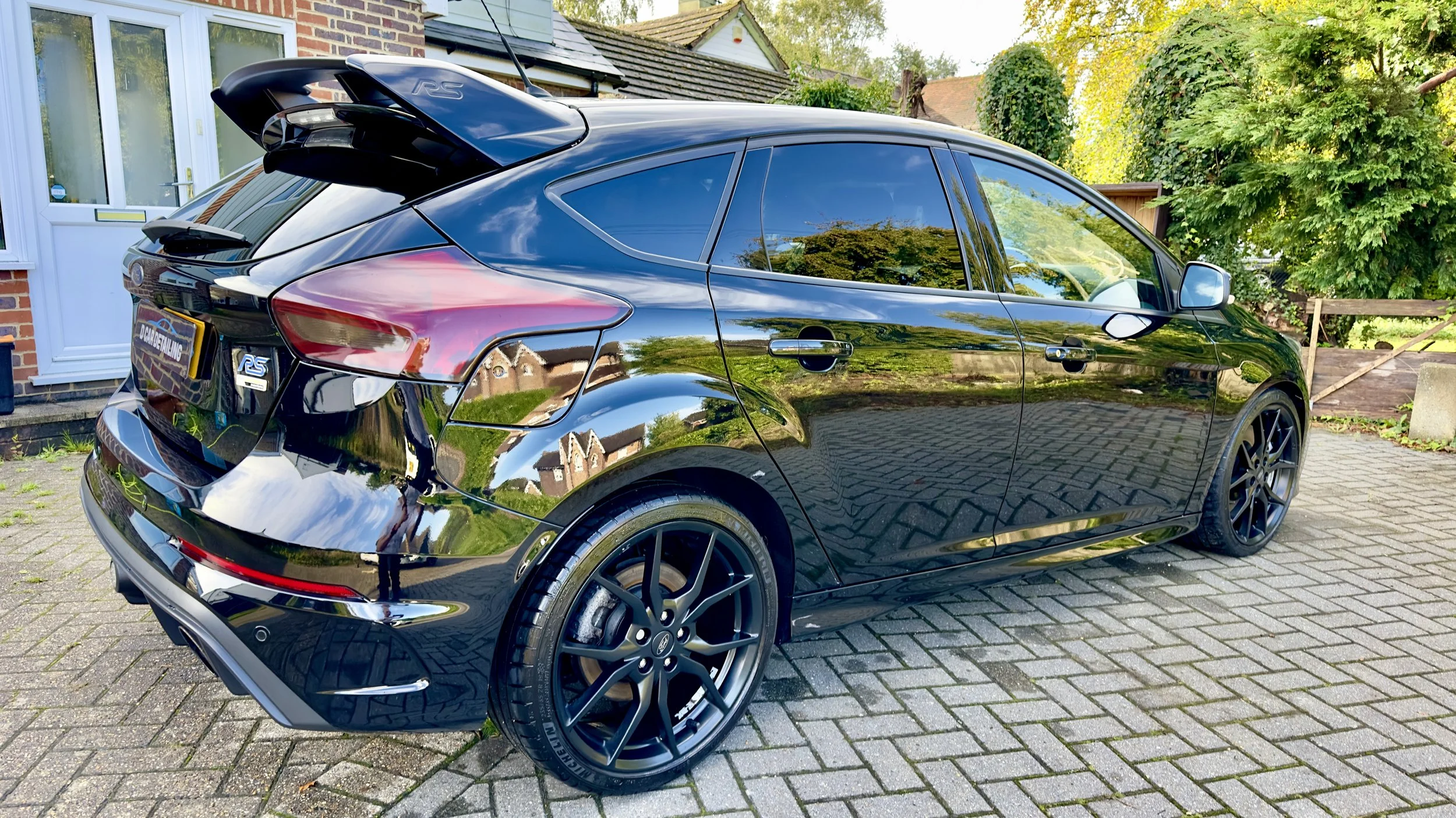 A black hatchback car parked on a brick driveway in front of a house surrounded by green trees and bushes.