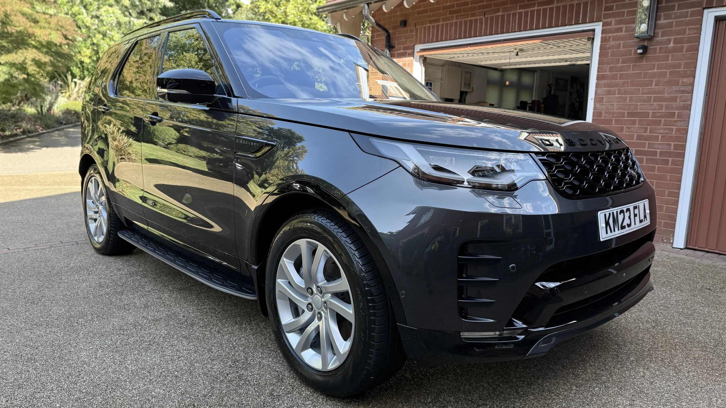 Black Land Rover Discovery Sport parked in driveway in front of a brick garage, reflecting the trees and sky on its shiny surface.