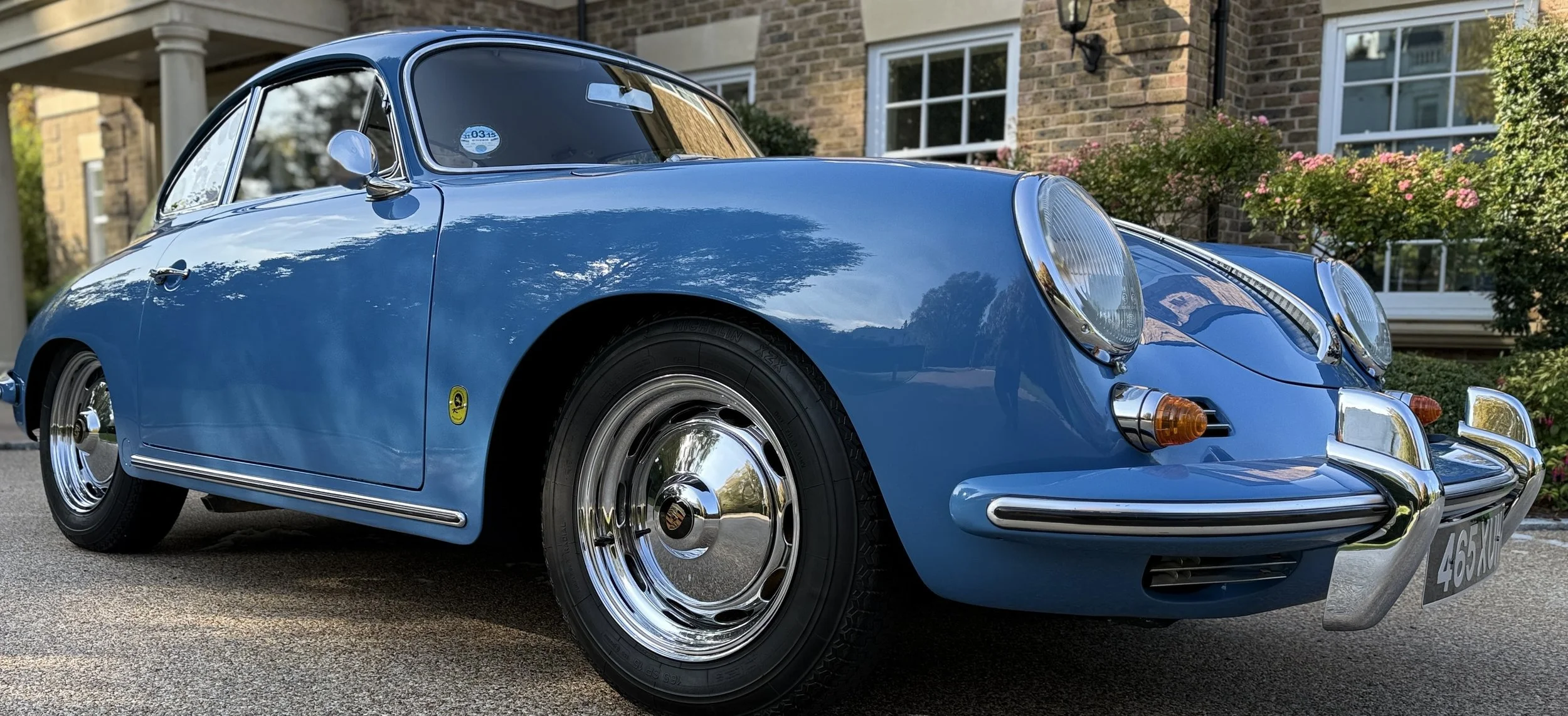 A classic blue Porsche 911 parked on a driveway in front of a brick house with garden flowers.