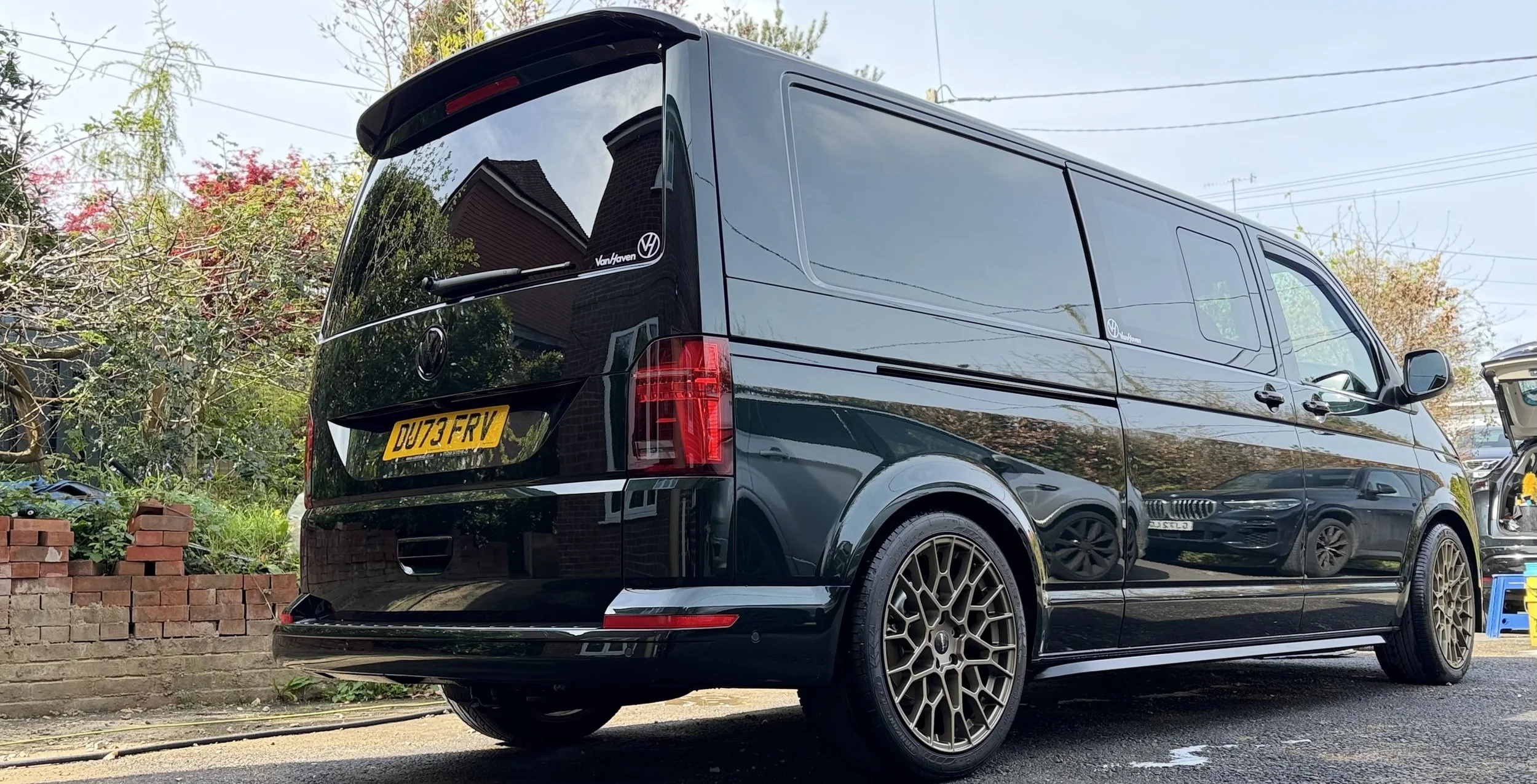 A black Volkswagen van parked on a residential street with trees, brick wall, and other parked cars in the background.