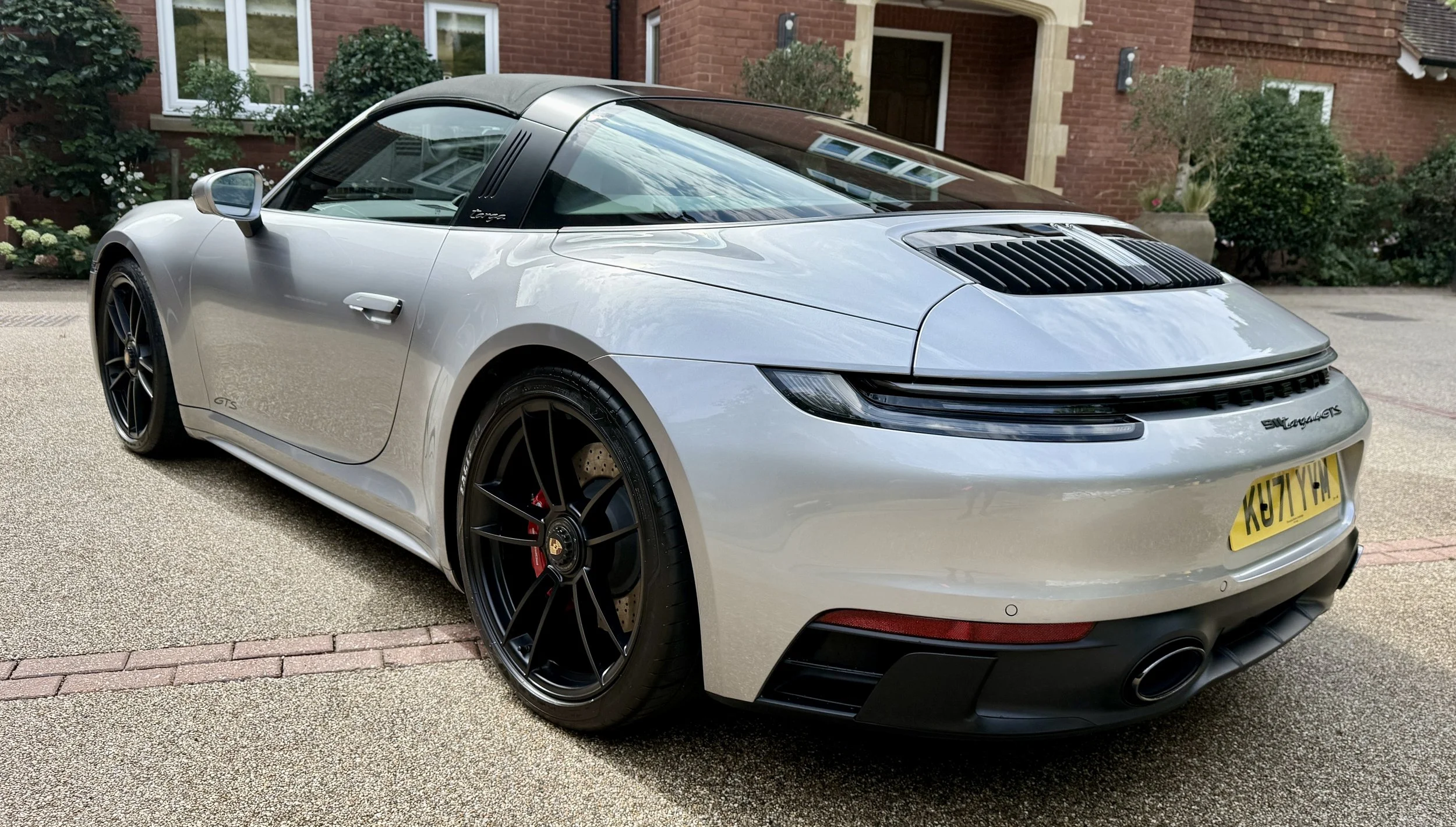 A silver Porsche 911 sports car parked in a residential driveway with a brick house and greenery in the background.