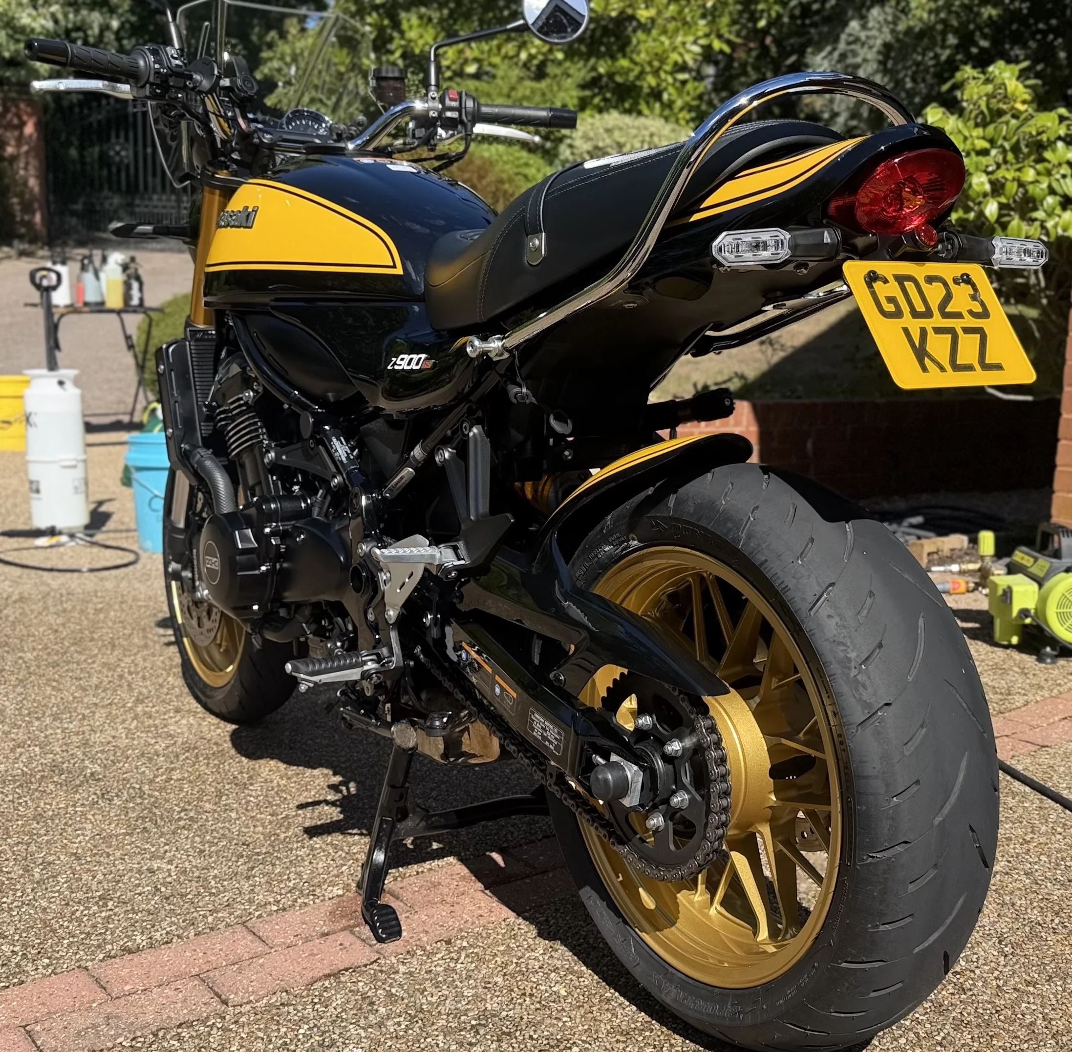 A black and gold Kawasaki motorcycle parked on a driveway with gravel surface, surrounded by greenery and tools in the background.