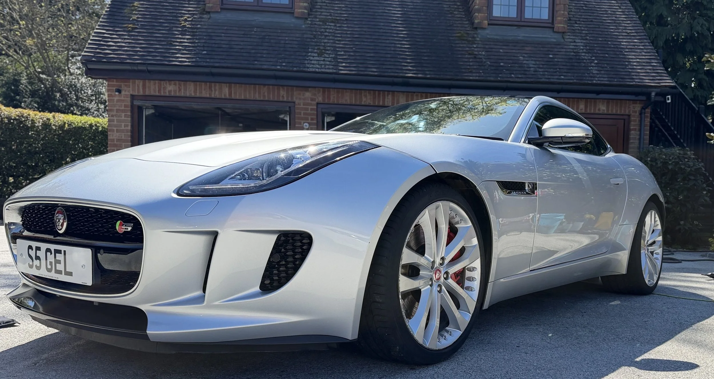 A silver Jaguar sports car parked in front of a brick house with a dark roof and large window.