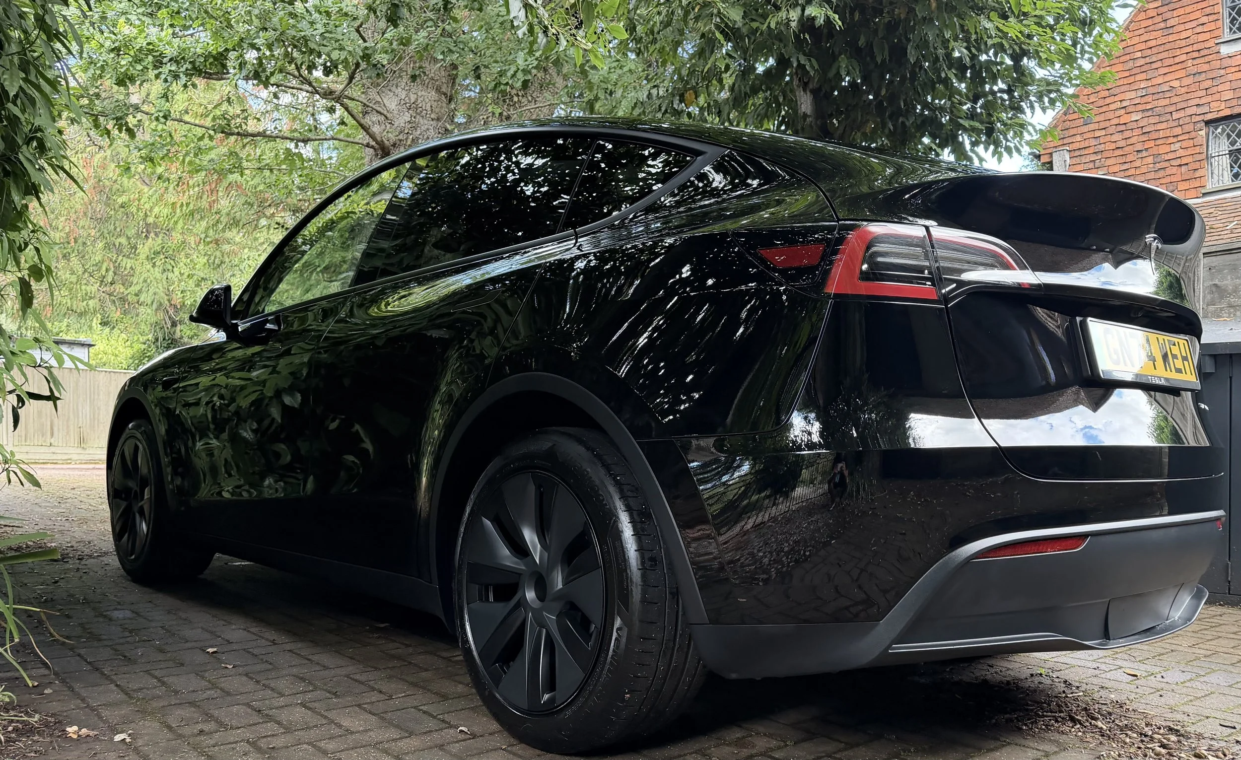 Black Tesla parked on a cobblestone driveway under trees with green leaves, a brick house with windows in the background.
