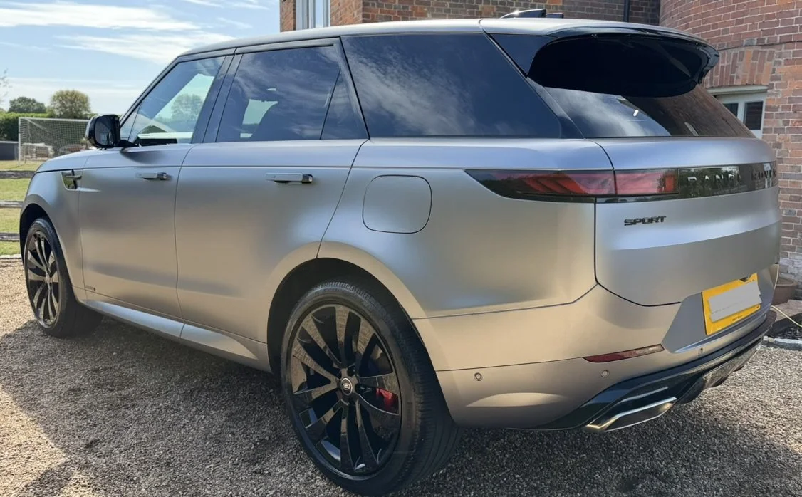 A silver Range Rover Sport parked on a gravel driveway near a brick house with a lawn and a fence in the background.
