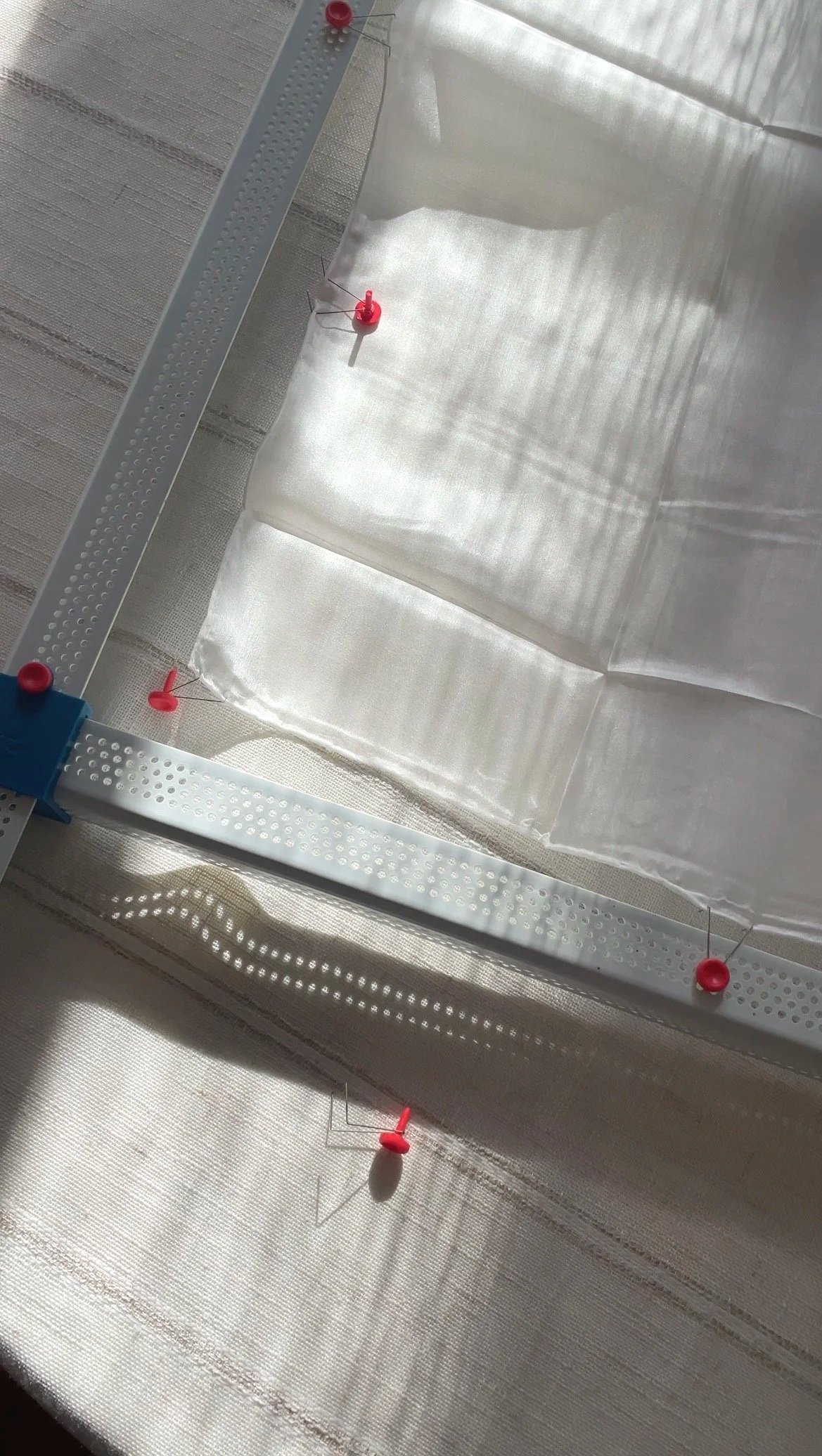 Close-up of a A partial view of a metal frame and red push pins securing sheer silk, sunlight creating shadows. First step in the batik creation process.