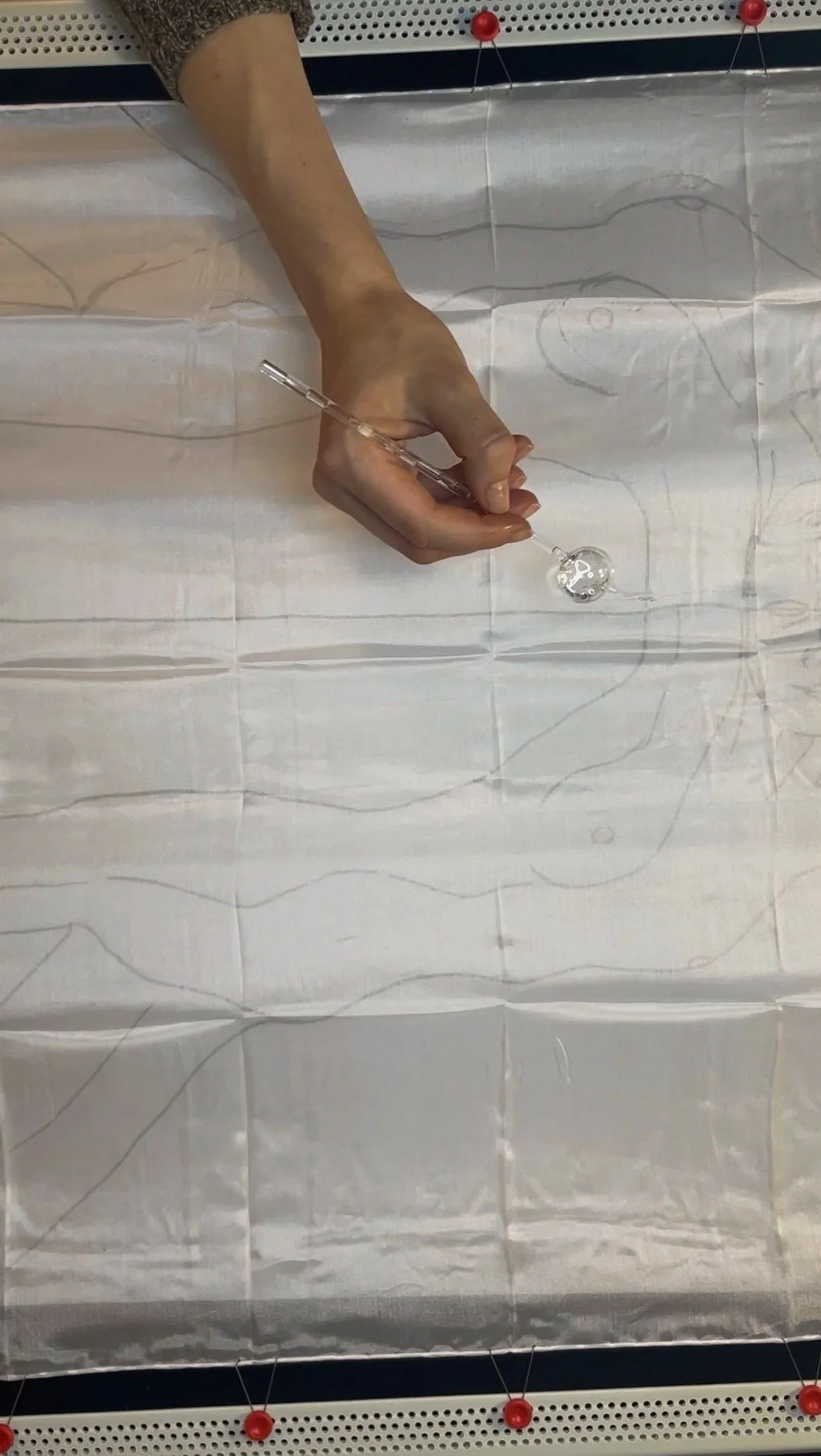 A person's hand holding a glass canting tool over a white sheer silk, secured with red pins on a metal frame.