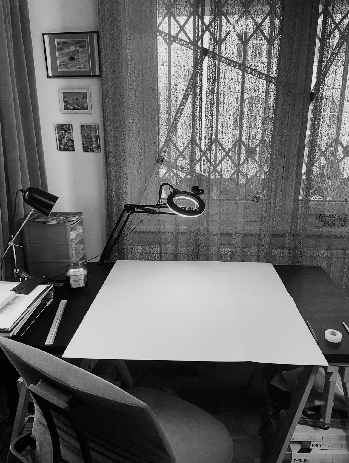 A studio workspace with a desk, chair, books, stack of notebooks, a ring light, and a lamp, in front of large window with lace curtains.