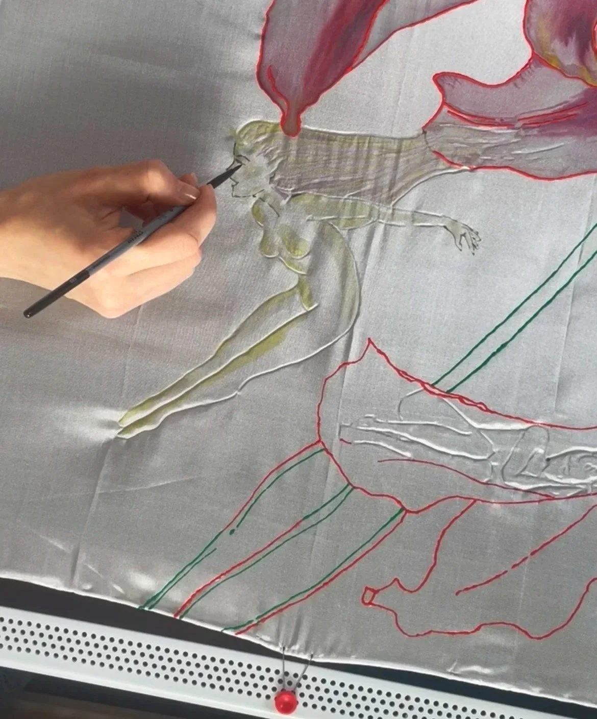 A person is drawing or colouring a detailed illustration of a fairy, sitting on a petal of a lilly, on silk with outlined sketches and coloured sections.