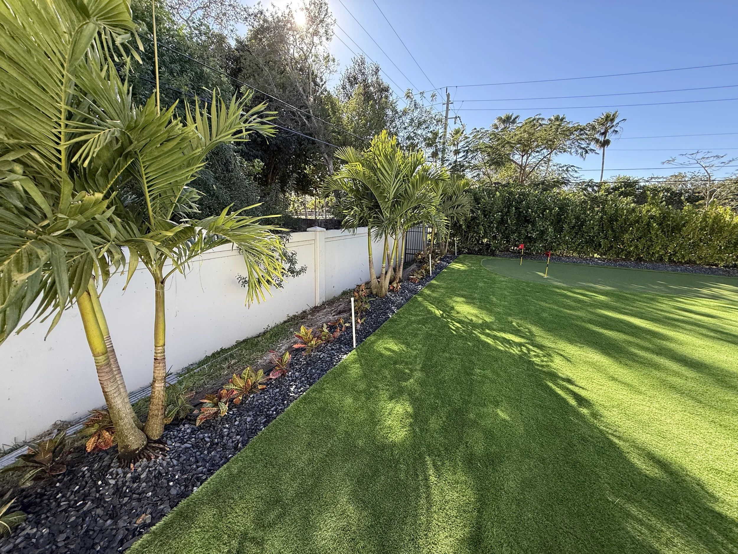 A backyard putting green with two small red golf flags, surrounded by tropical plants and a white fence, under a clear blue sky.