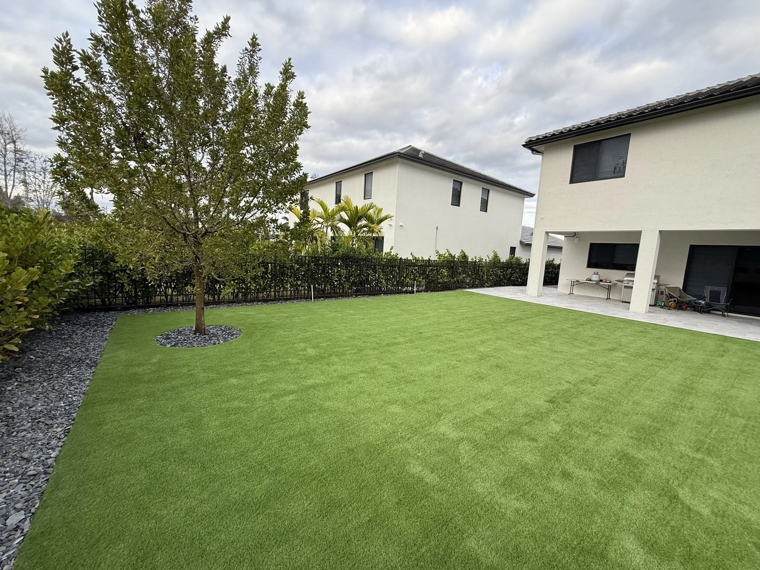 Backyard with artificial green grass, a tree with mulch around its base, a black metal fence, and two white modern houses with black windows and covered patios.