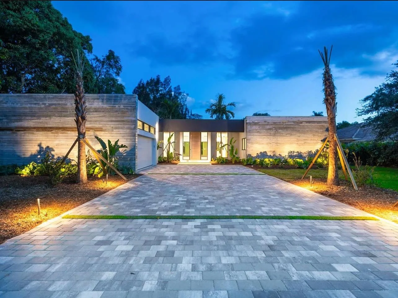 Modern house with a large driveway, palm trees, and landscape lighting at dusk.