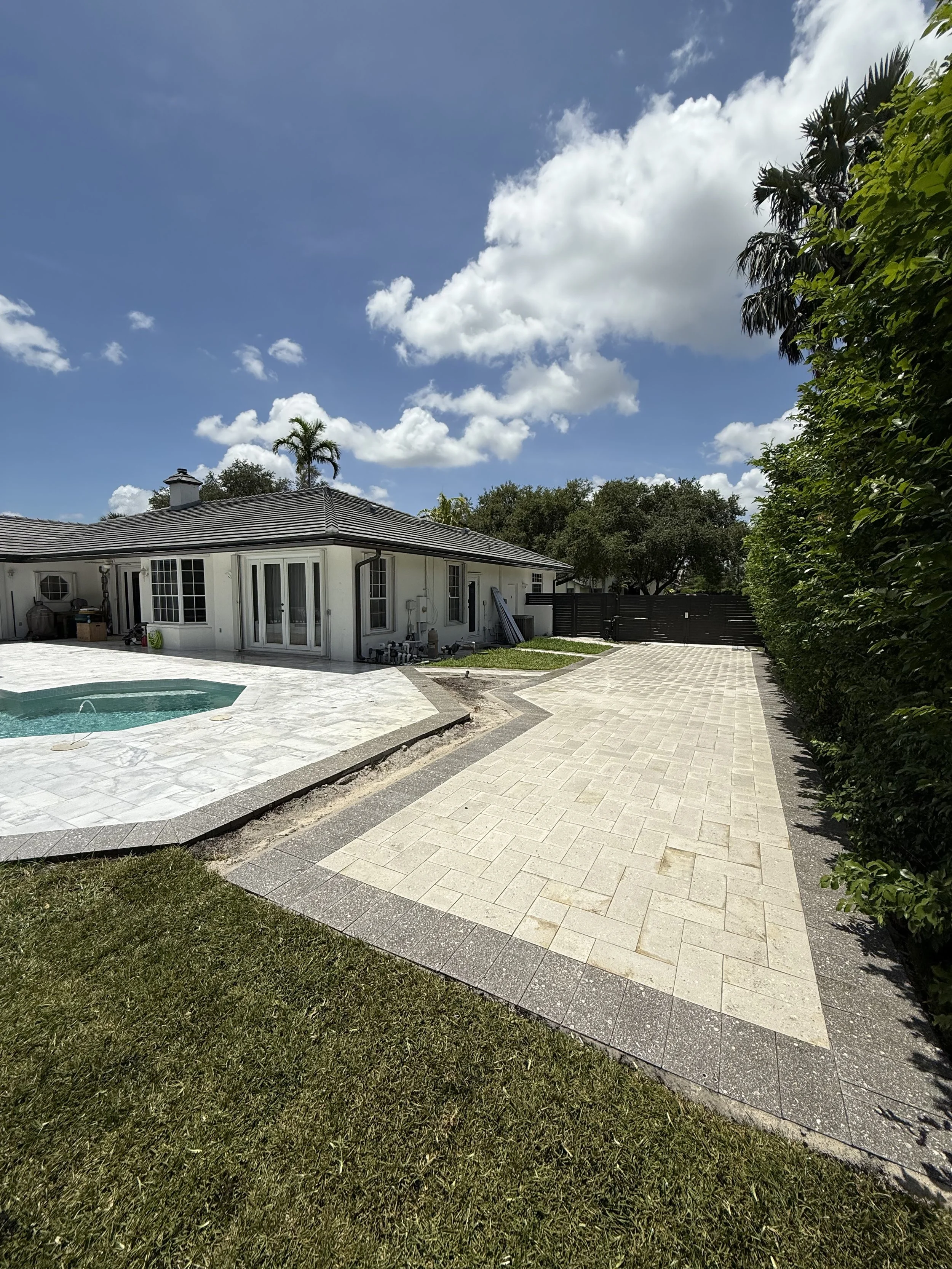 A backyard with a swimming pool, paved walkway, and a white house under a partly cloudy sky.