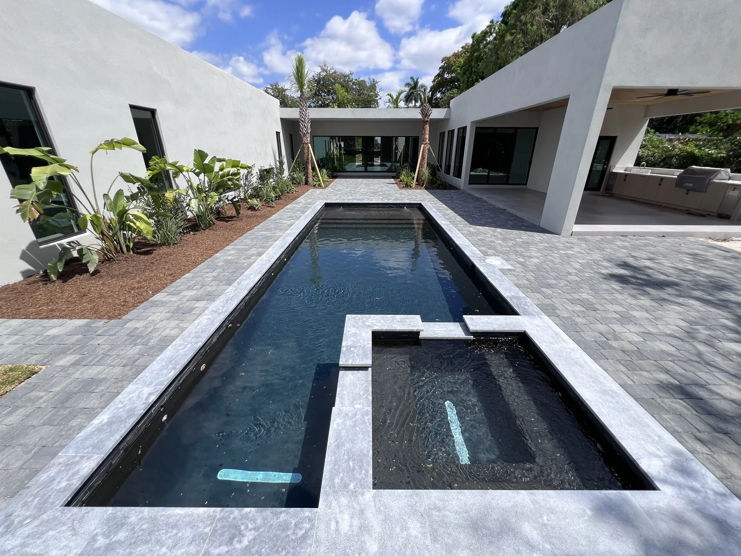 Modern backyard with rectangular swimming pool featuring a built-in hot tub, surrounded by gray stone pavers, with planters and two palm trees, adjacent outdoor kitchen and covered patio area.