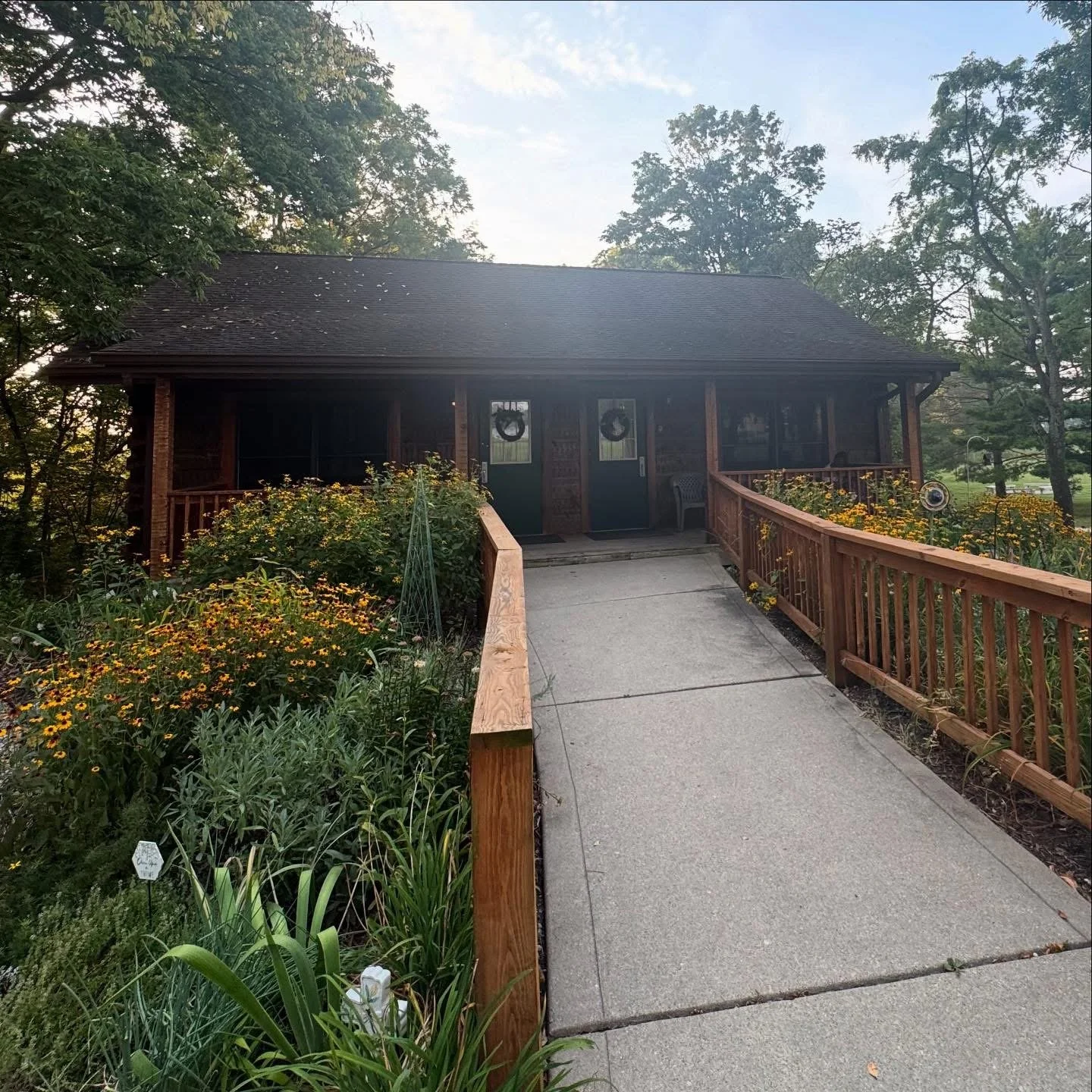 A single-story house with a dark roof, front porch, and glass doors decorated with wreaths. A concrete walkway with wooden railings leads up to the house, surrounded by flowers and greenery.