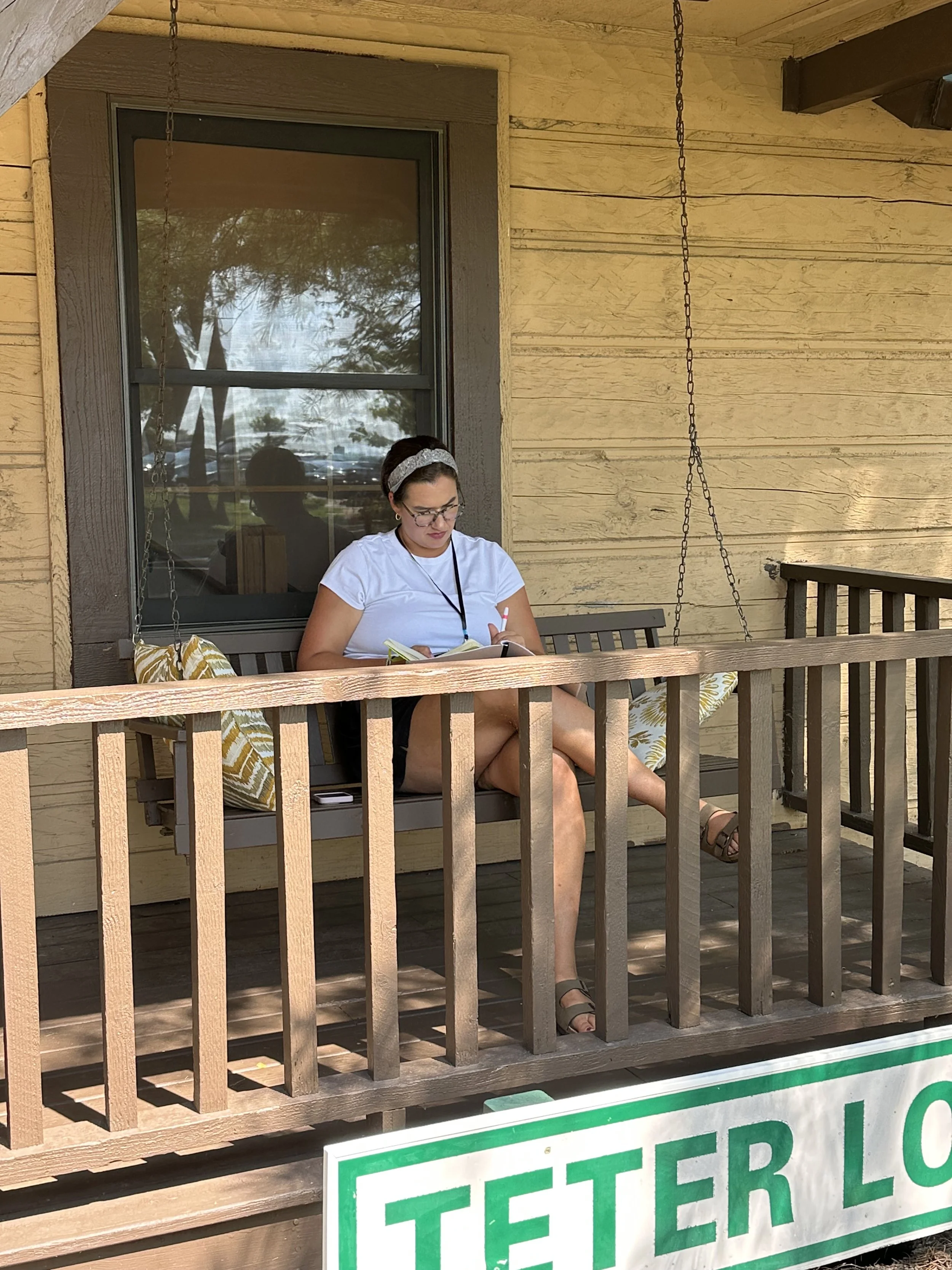A woman sitting on a porch swing on a wooden porch, reading a book, with a window behind her reflecting trees and sky, and a sign that reads 'PETER LO' at the bottom of the image.