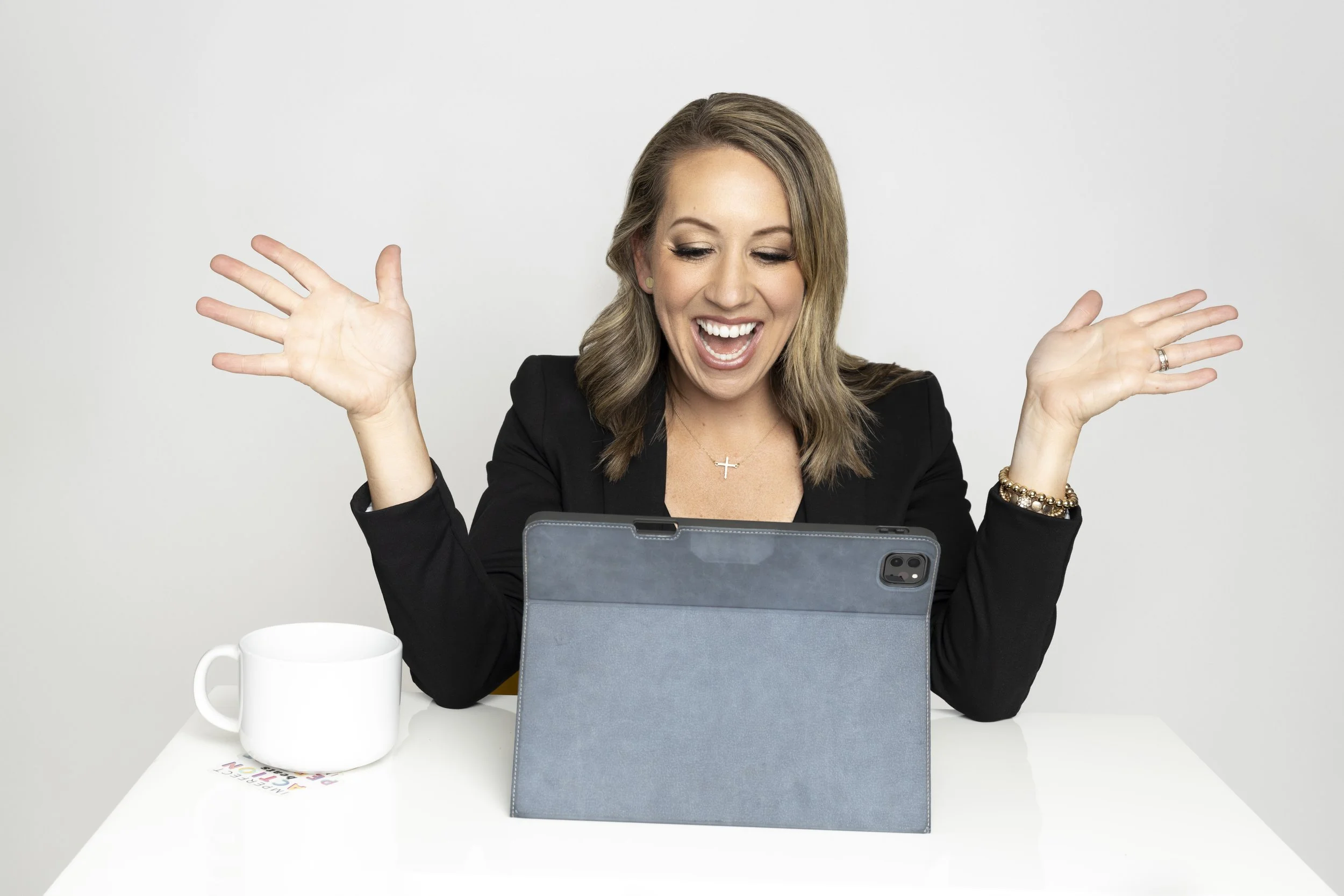 A woman in a black blazer excitedly looks at a tablet on a table, with her hands raised. There is a white mug and a colorful paper on the table.