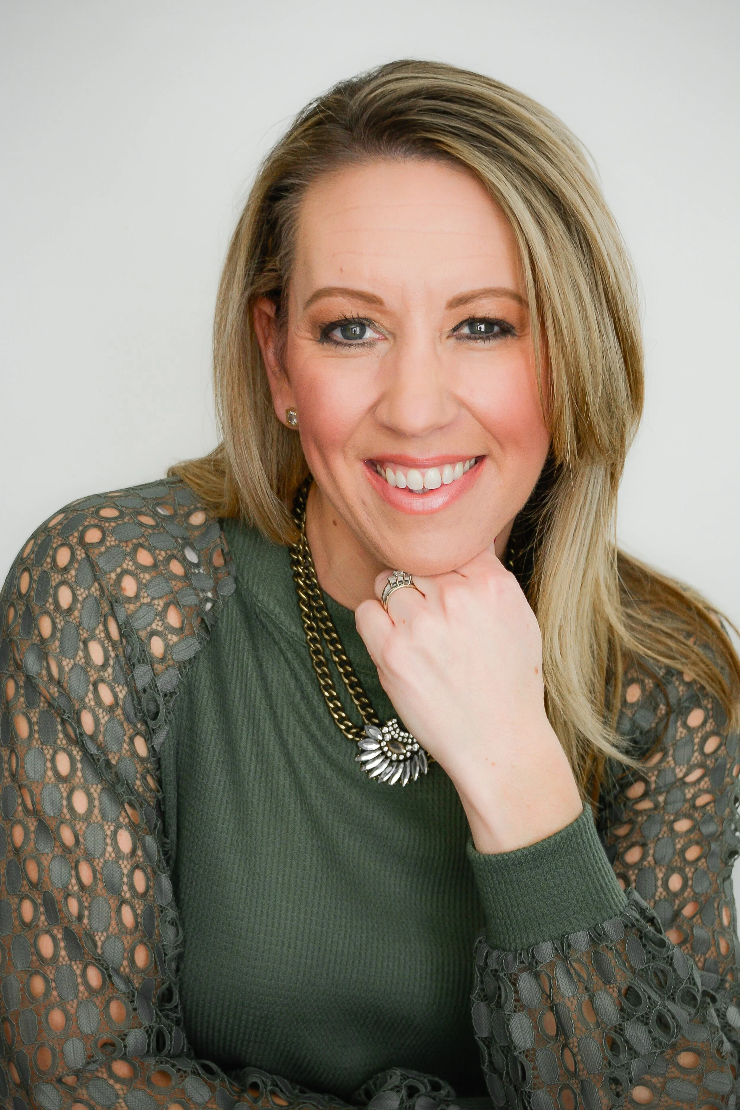A woman with blonde hair smiling, resting her chin on her hand, wearing a green top with lace sleeves and a statement necklace.