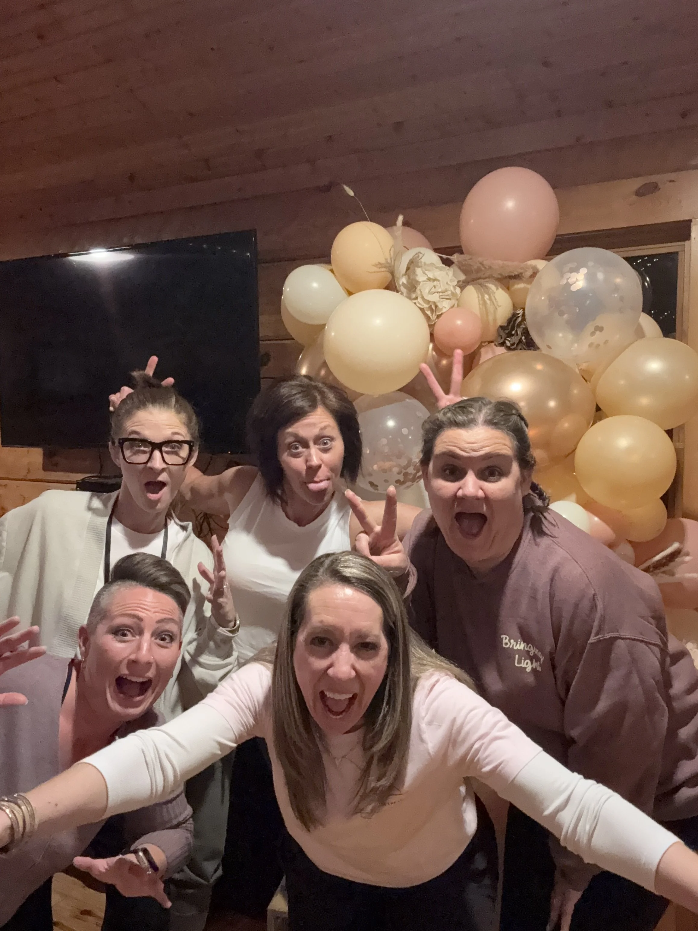 Group of five women celebrating at a party with a balloon arrangement behind them. They are making playful faces and hand gestures, with some giving bunny ears behind others' heads, in a cozy wooden room.
