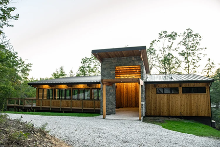 Modern house with a wooden and stone exterior, large windows, and a metal roof, surrounded by trees.